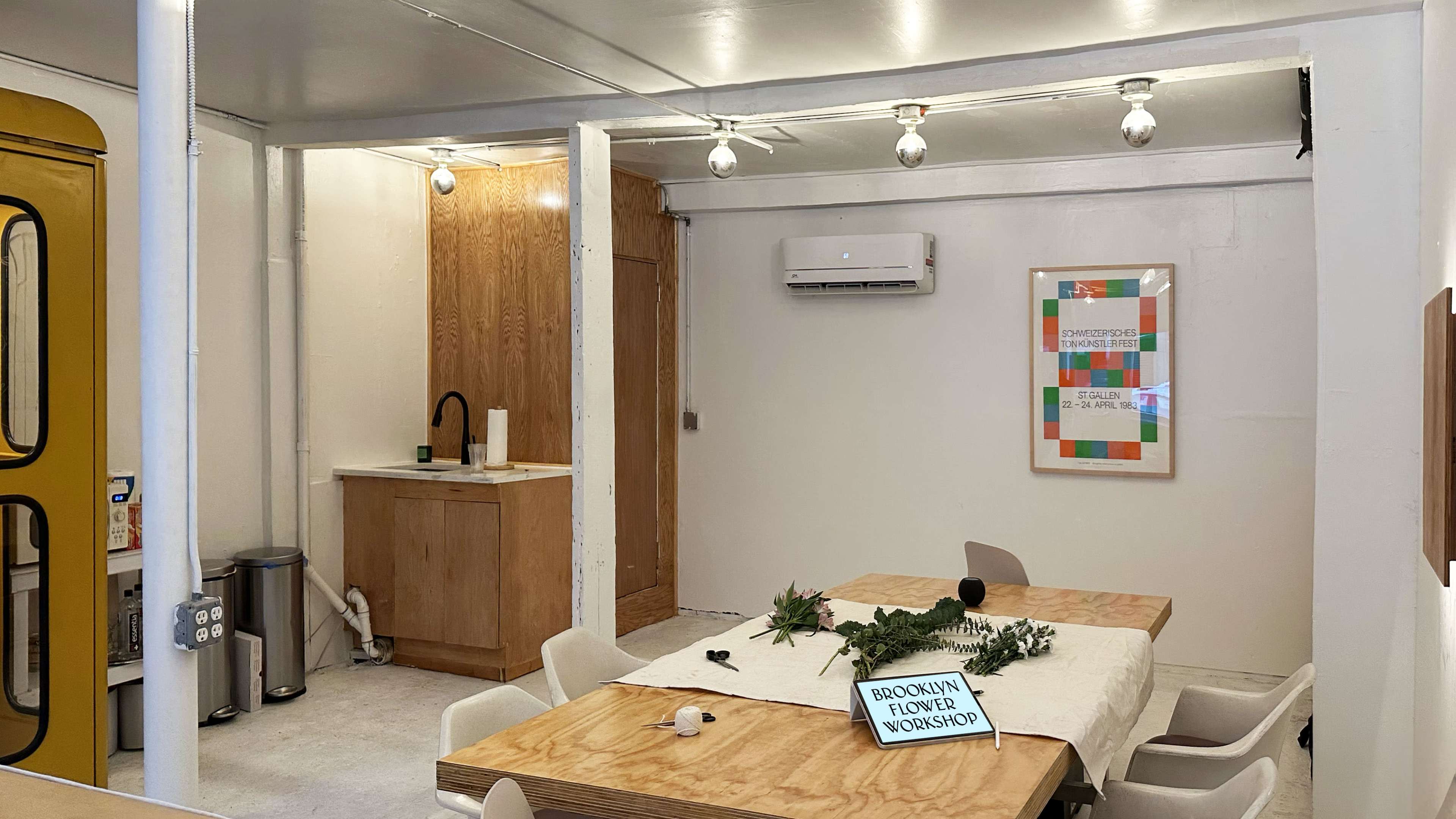 A bright, modern workshop space with a wooden table, a kitchen area in the background, and a sign indicating a Brooklyn flower workshop.