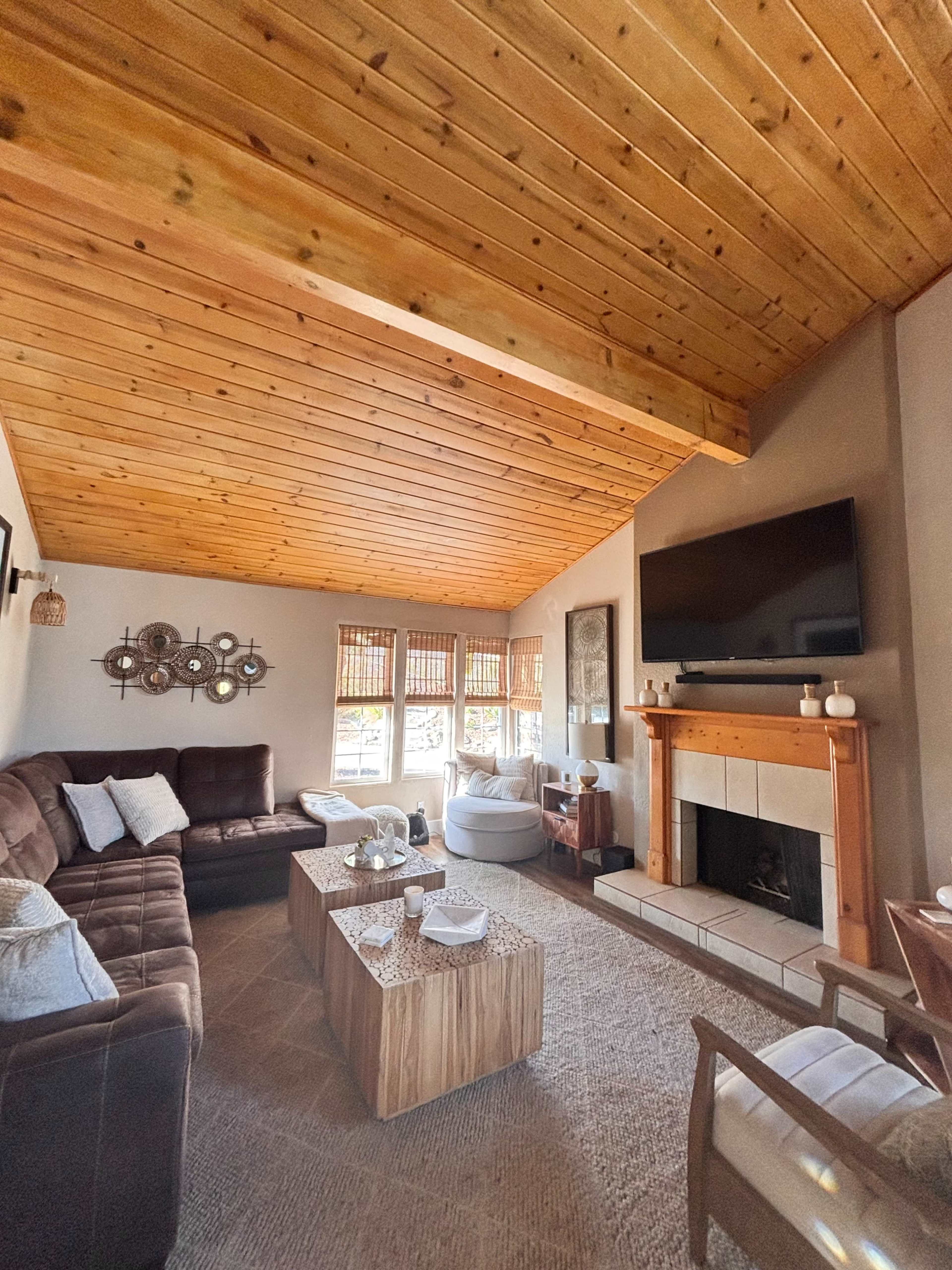 The image shows a cozy living room with a wooden ceiling, a brown sectional sofa, a stone fireplace, and large windows with woven shades.