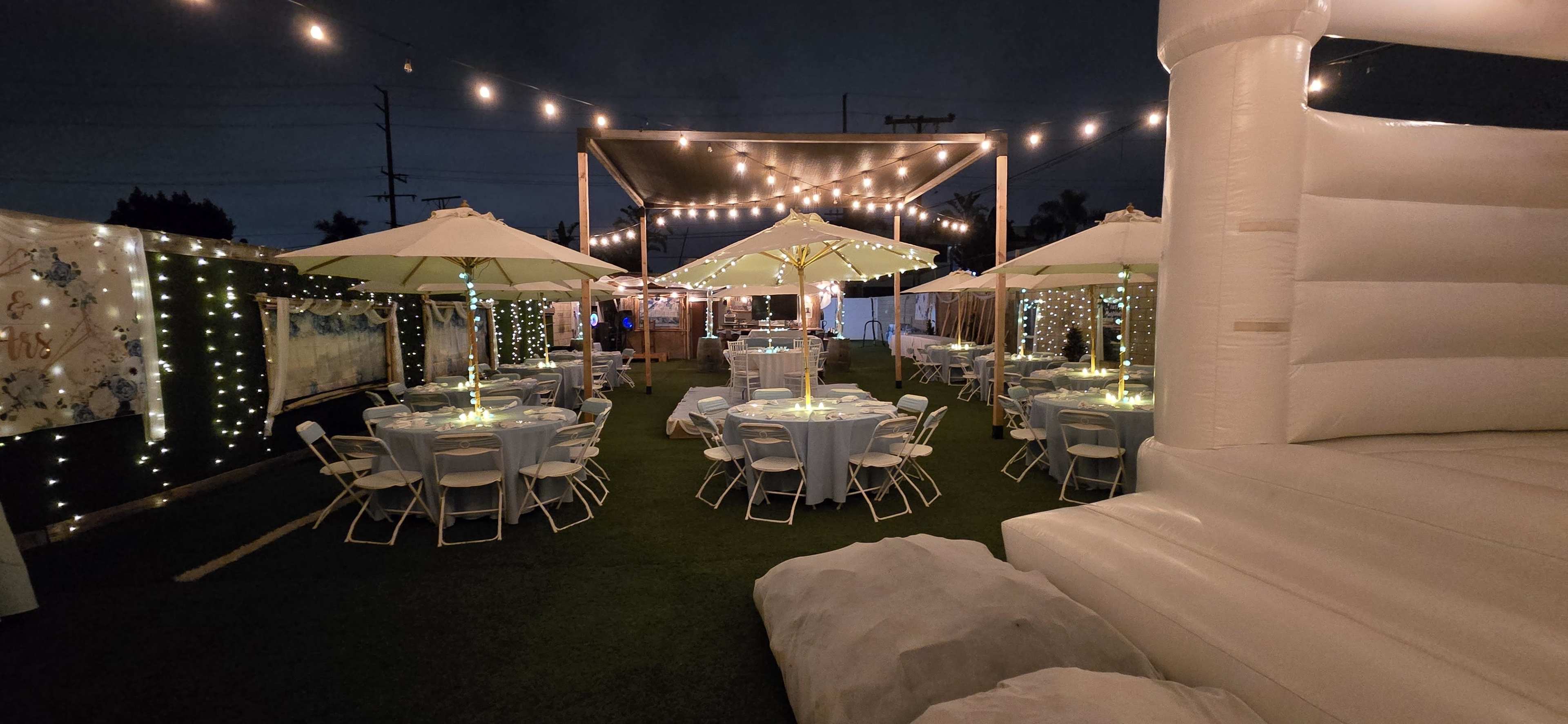 A decorated outdoor event space at night with several tables, umbrellas, and string lights illuminating the area.