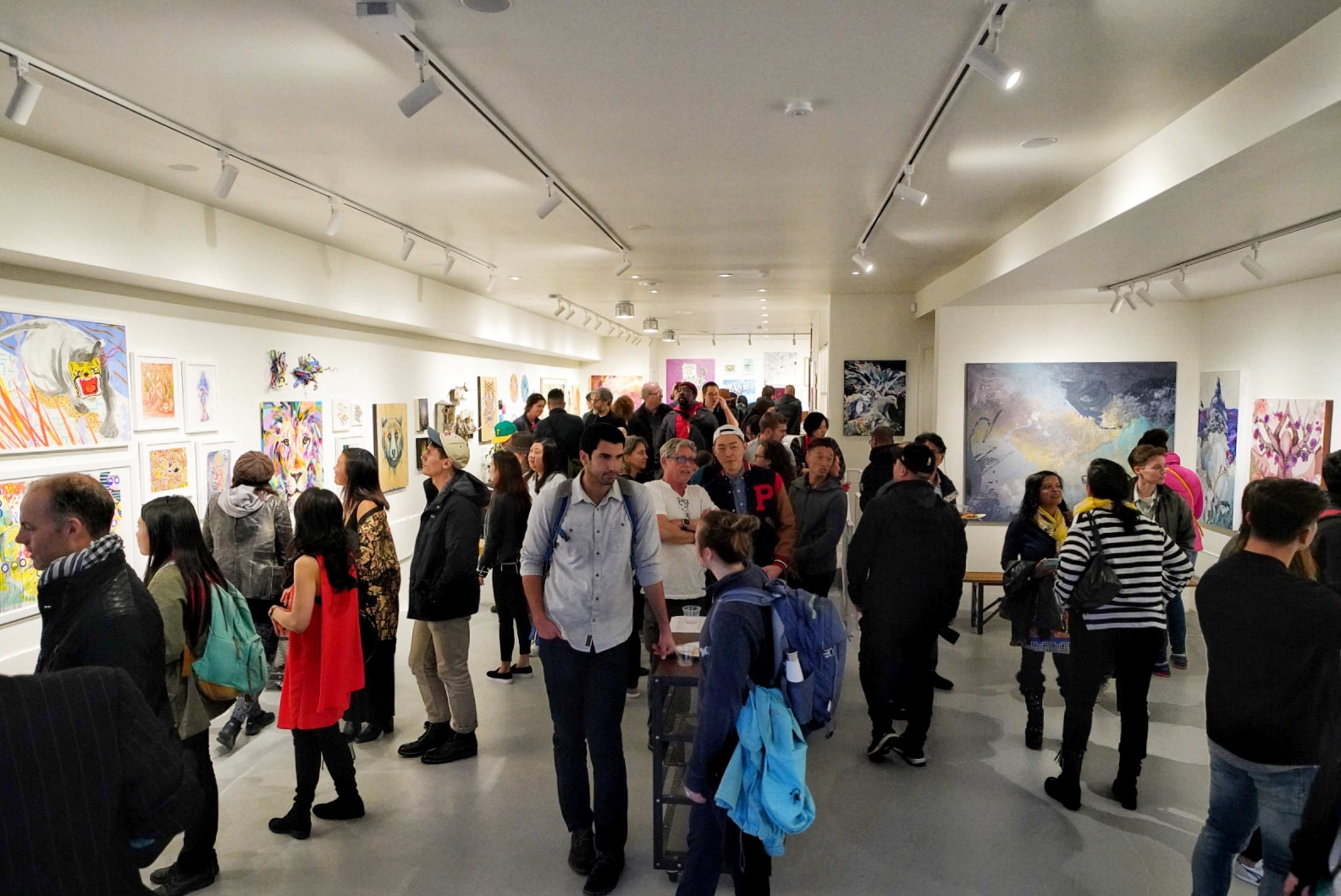 A crowded art gallery features people viewing various artworks displayed on the walls.