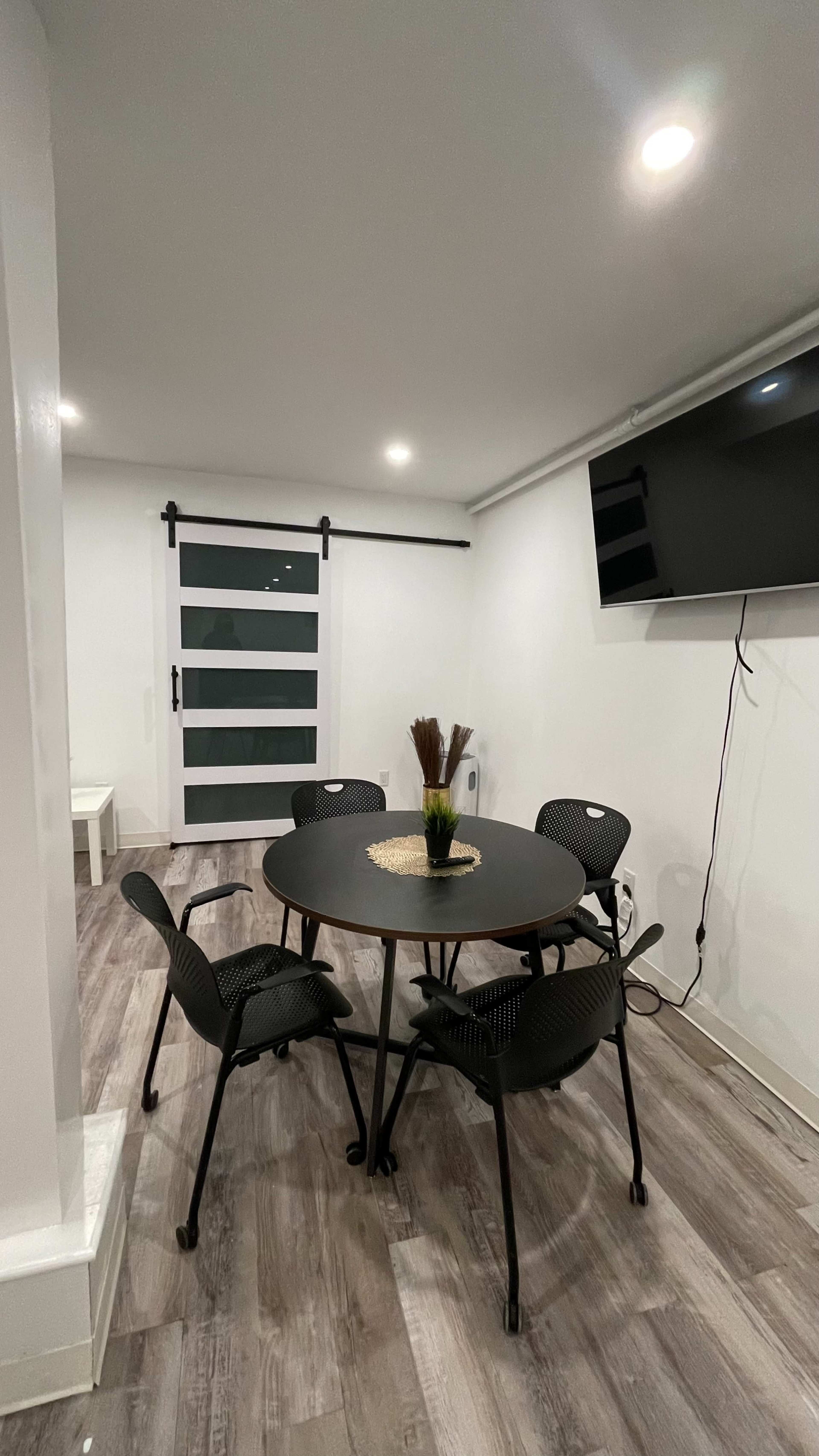 A modern room with a round black table and five black chairs, featuring a sliding glass door and a mounted television on the wall.