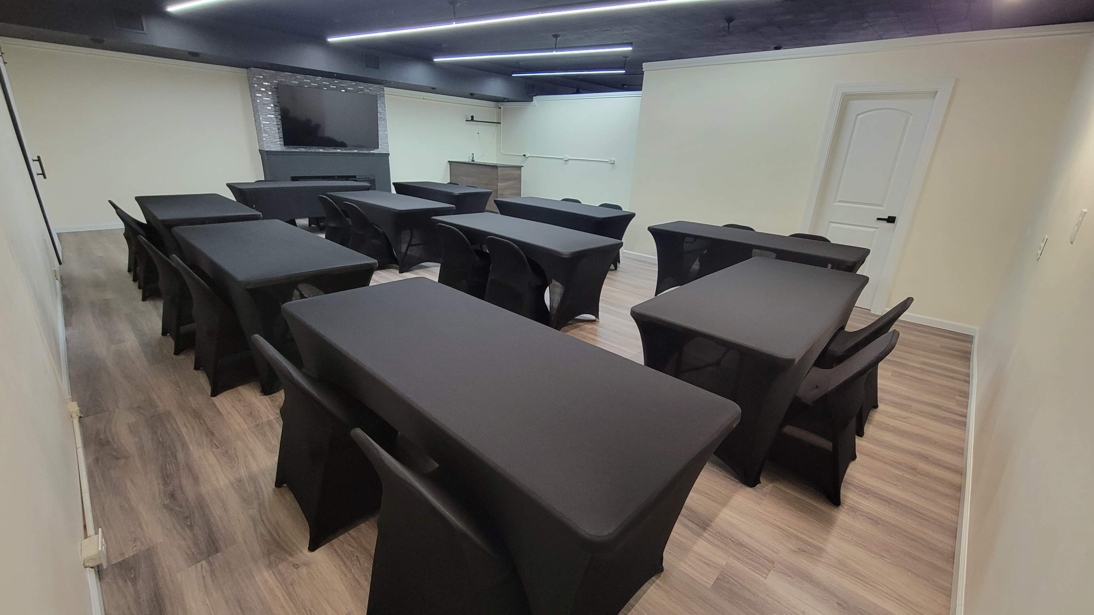 A conference room arranged with multiple tables covered in black cloth and chairs, with a television and a corner kitchenette visible in the background.