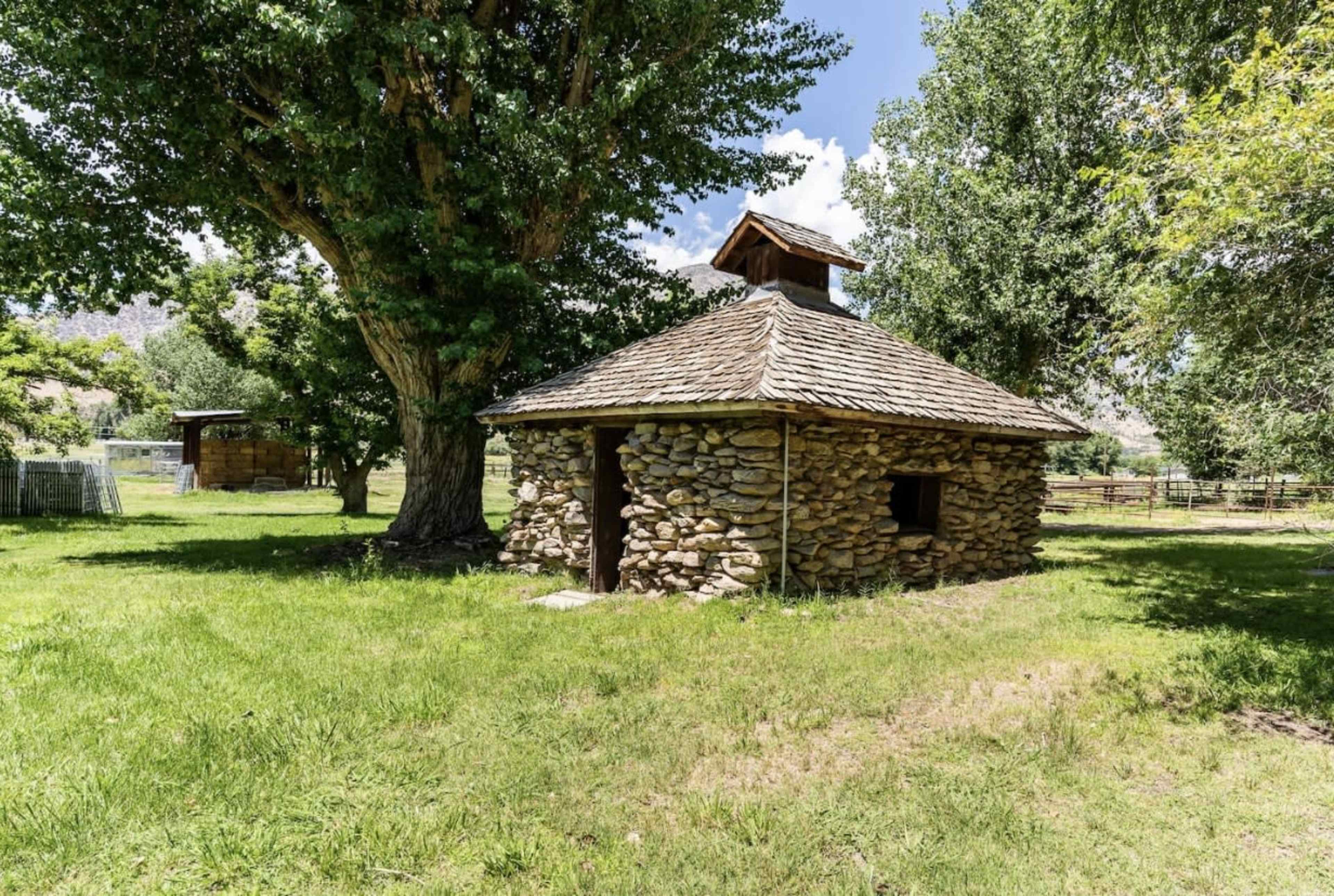 A stone building with a wooden roof stands in a grassy area surrounded by trees.