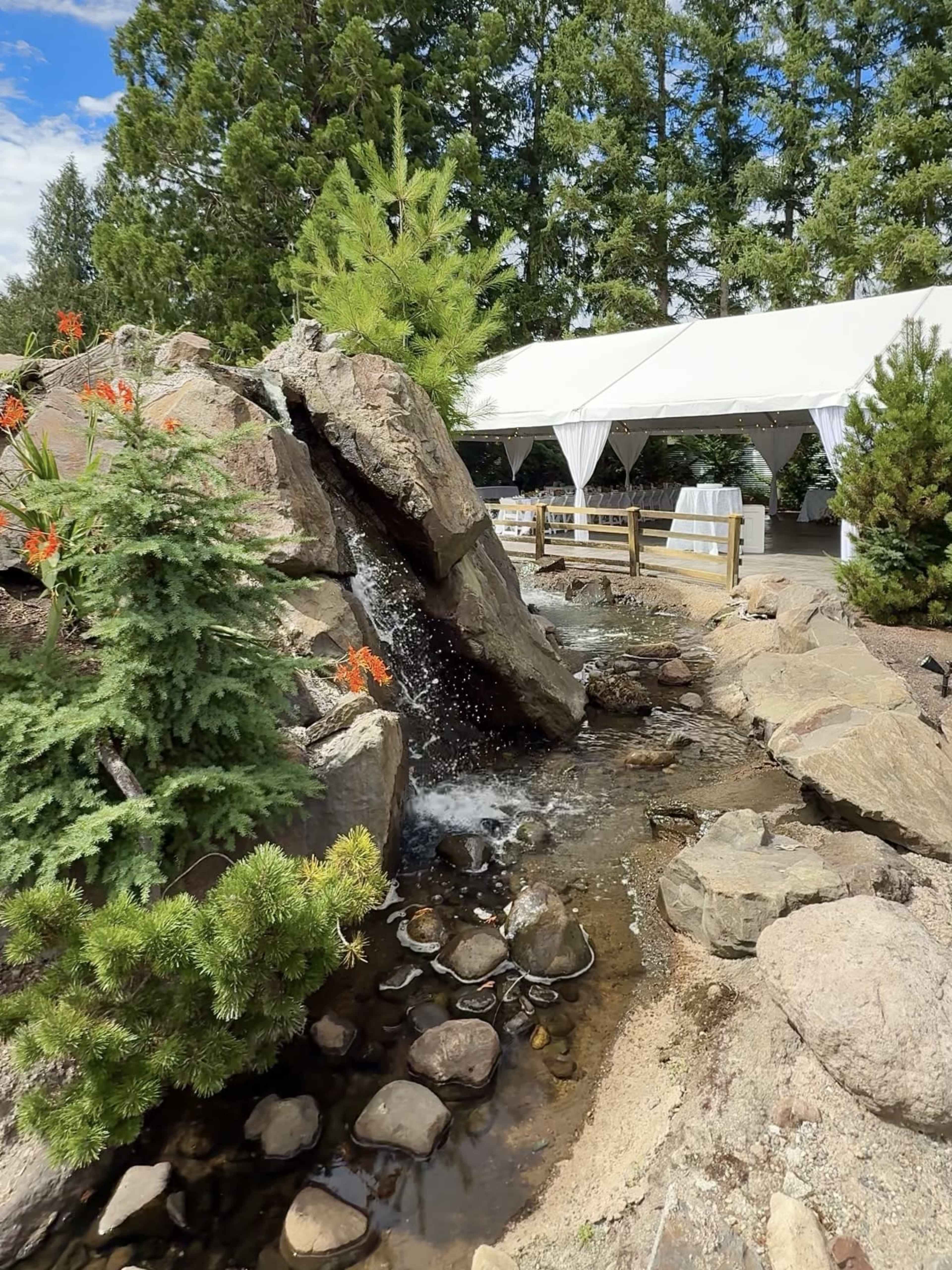 A small waterfall flows over rocks into a landscaped area with flowers and a white tent in the background.