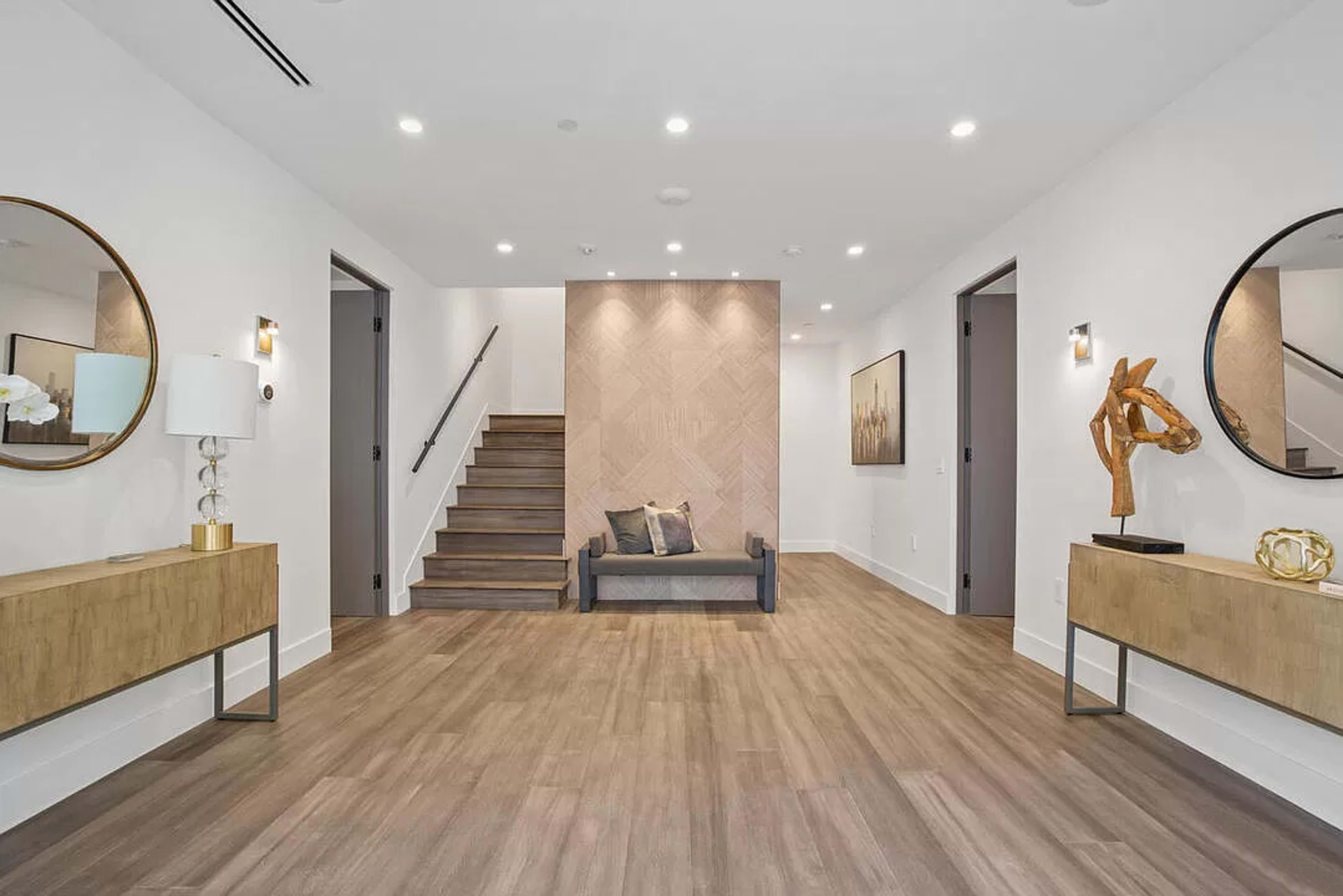 A modern foyer features wooden flooring, a staircase, two round mirrors, and contemporary decor elements, including a sculpture and side tables.