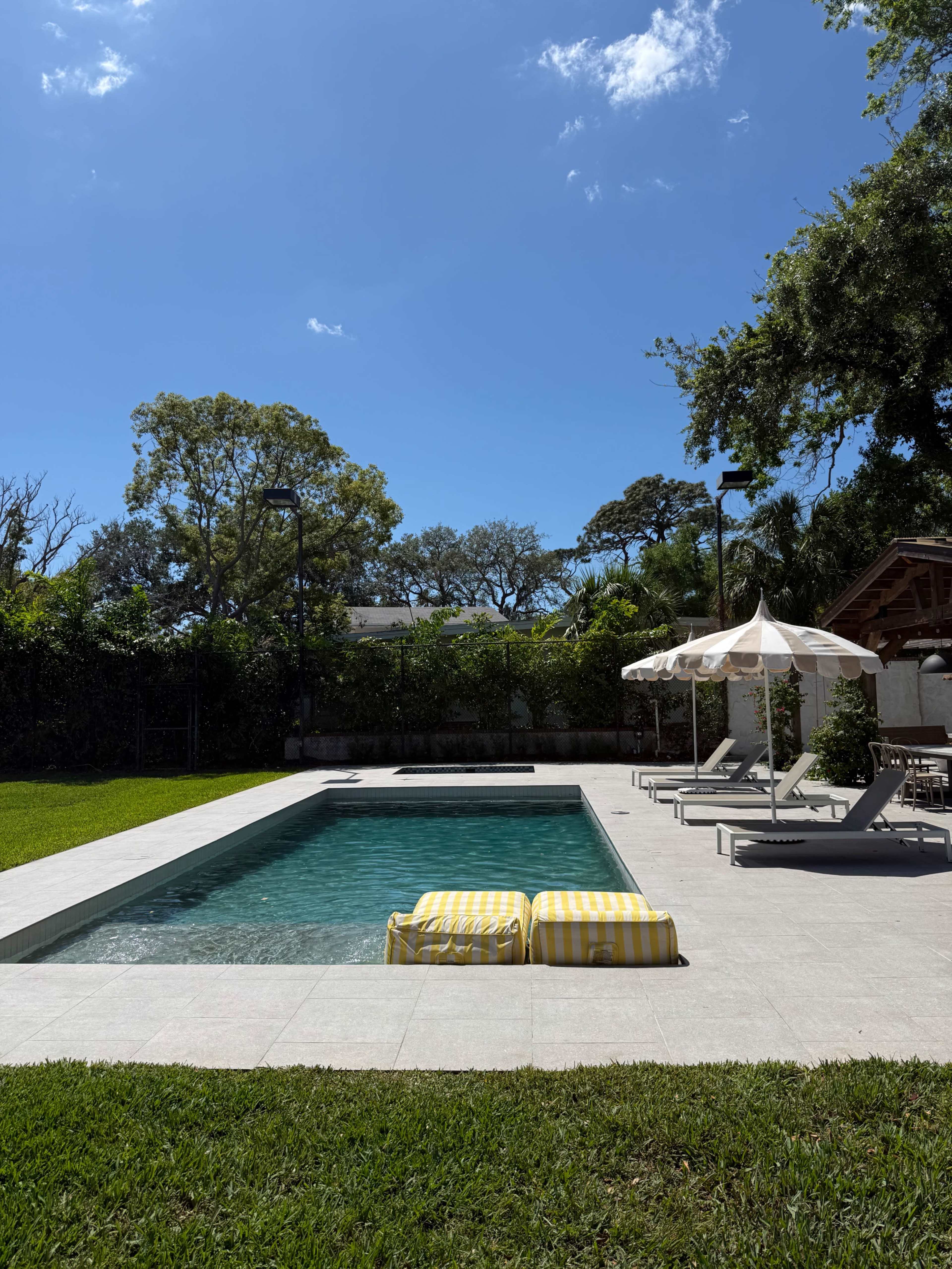 The image shows a rectangular swimming pool surrounded by lounge chairs and yellow-striped floats, with greenery and a clear blue sky in the background.