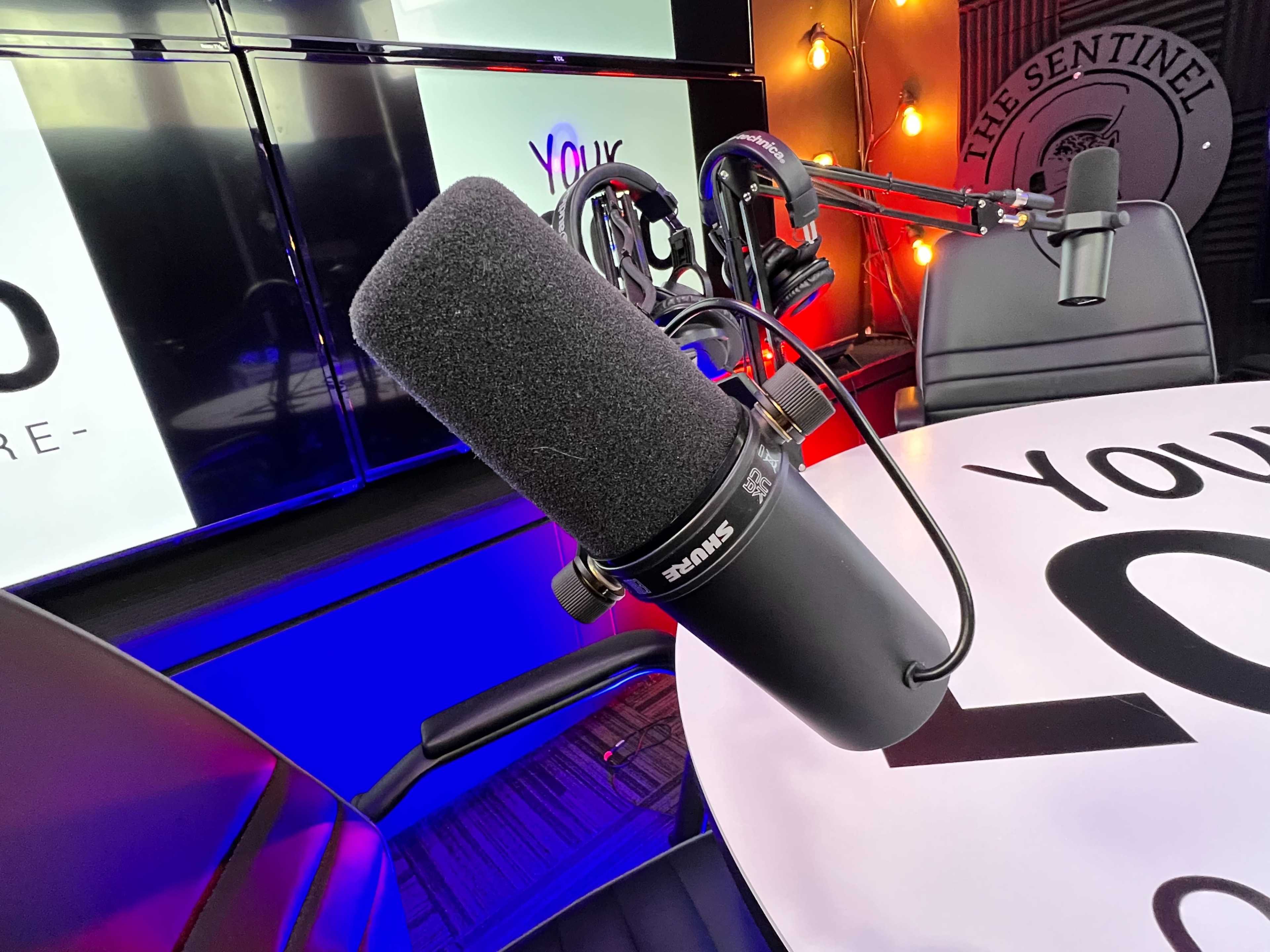 A close-up of a microphone on a table in a studio with colorful lighting and screens in the background.