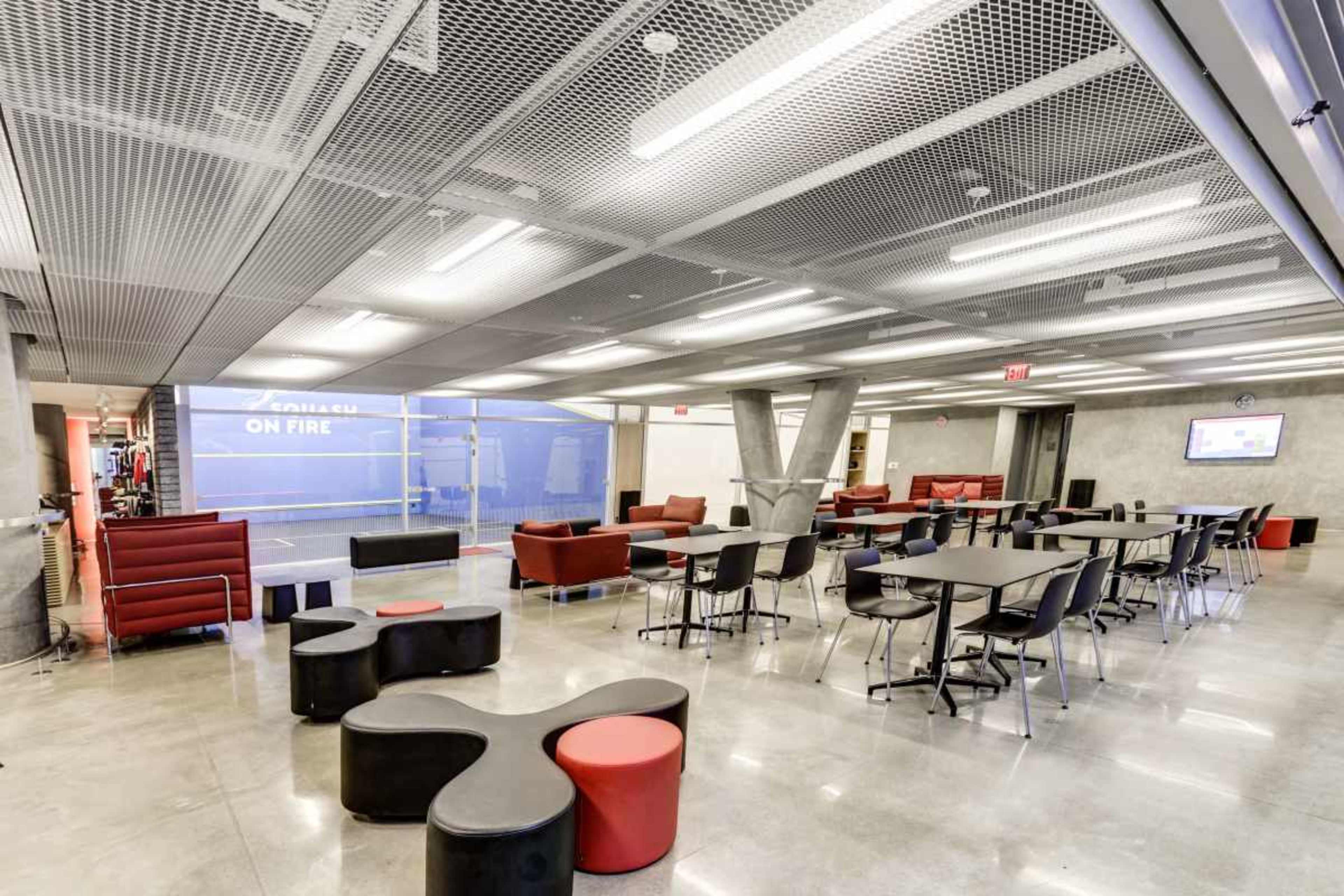 The image shows a modern, open-plan lounge area with red and black furniture, including sofas and tables, under a metal mesh ceiling with bright lighting.
