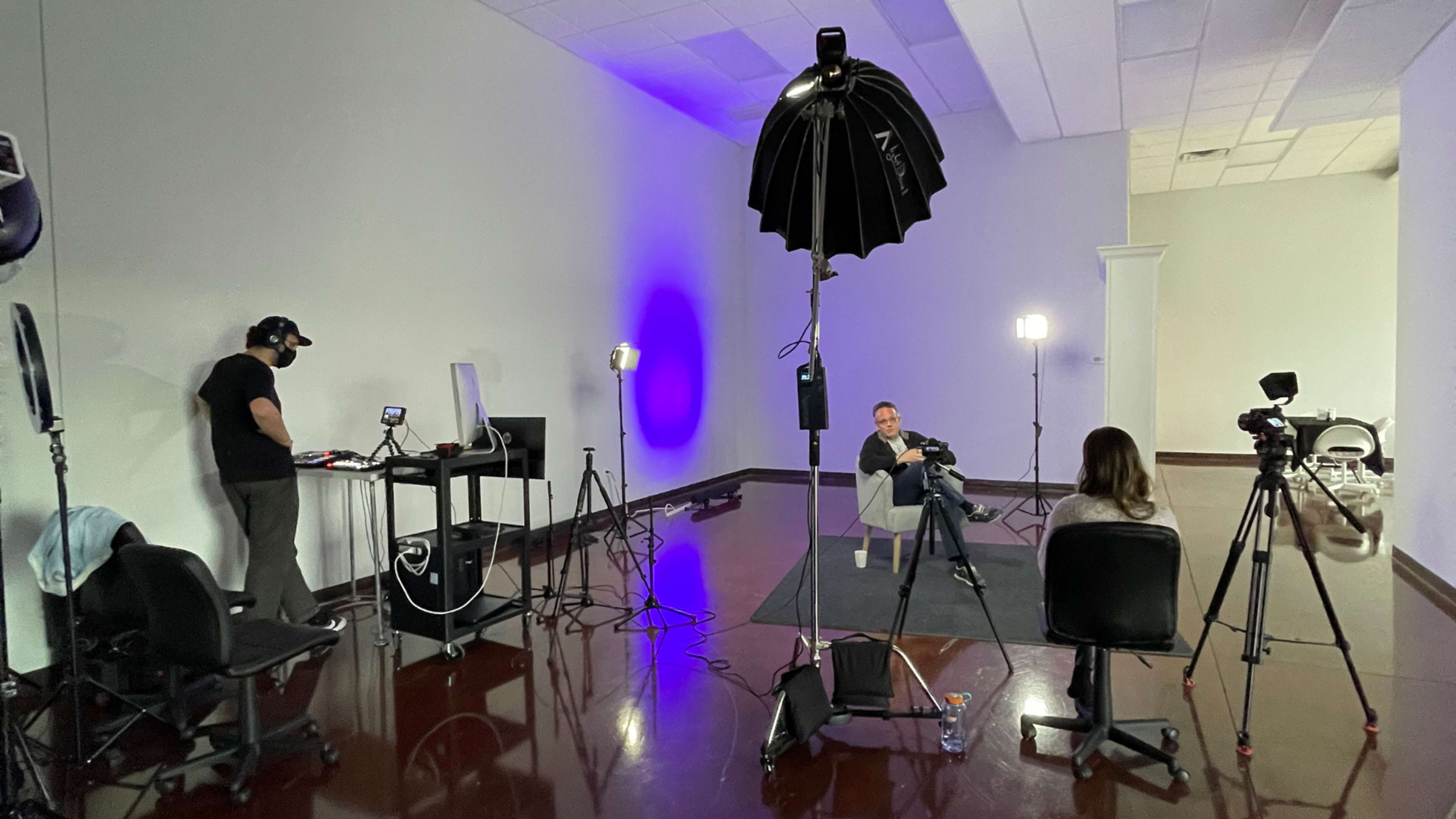 A video recording setup features a seated man being interviewed, flanked by cameras and studio lights, with a technician operating equipment in the background.