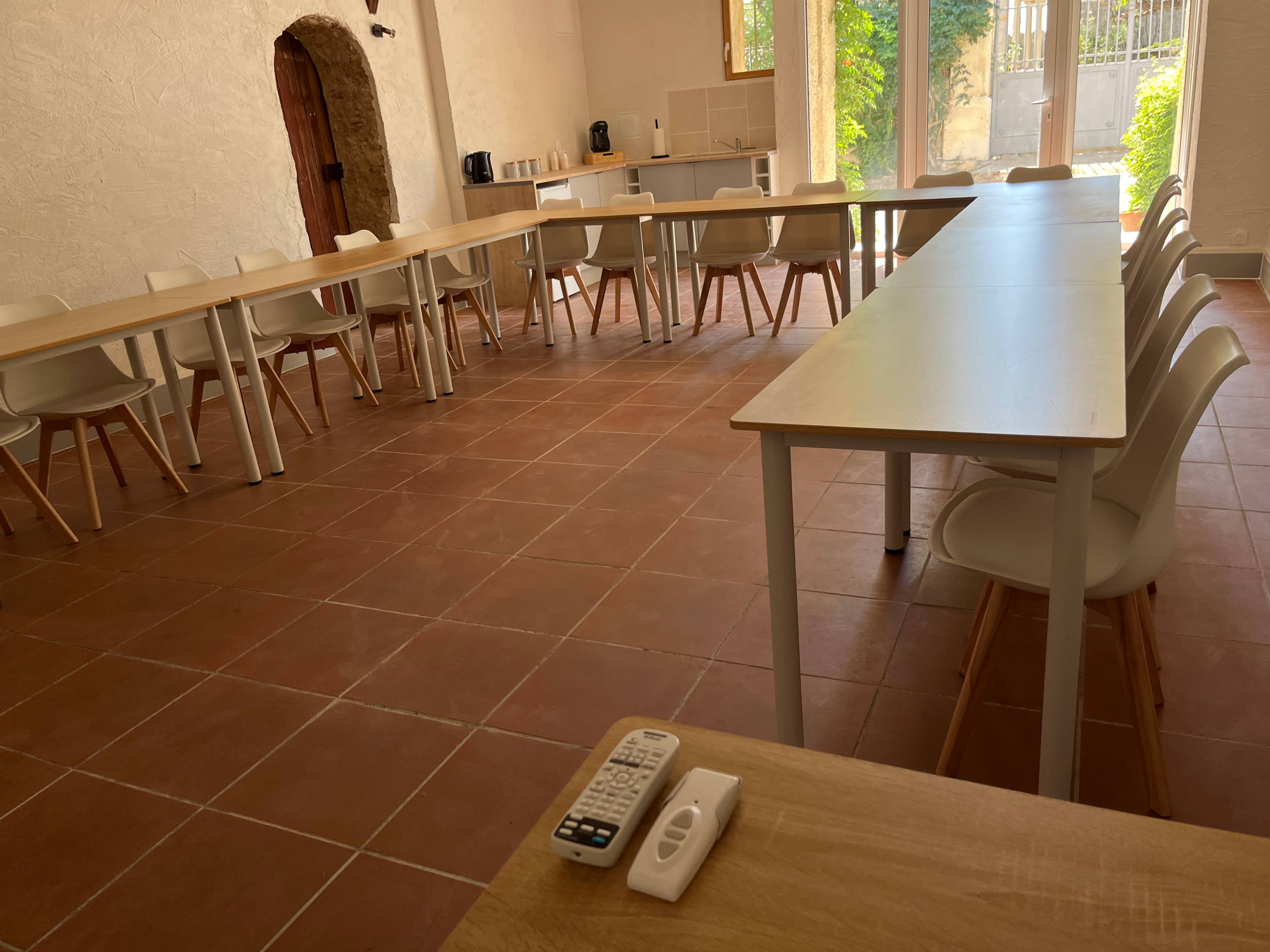 The image shows a spacious room with a tiled floor, featuring several tables and chairs arranged in a dining or meeting setup, along with a remote control and a small device on a table in the foreground.