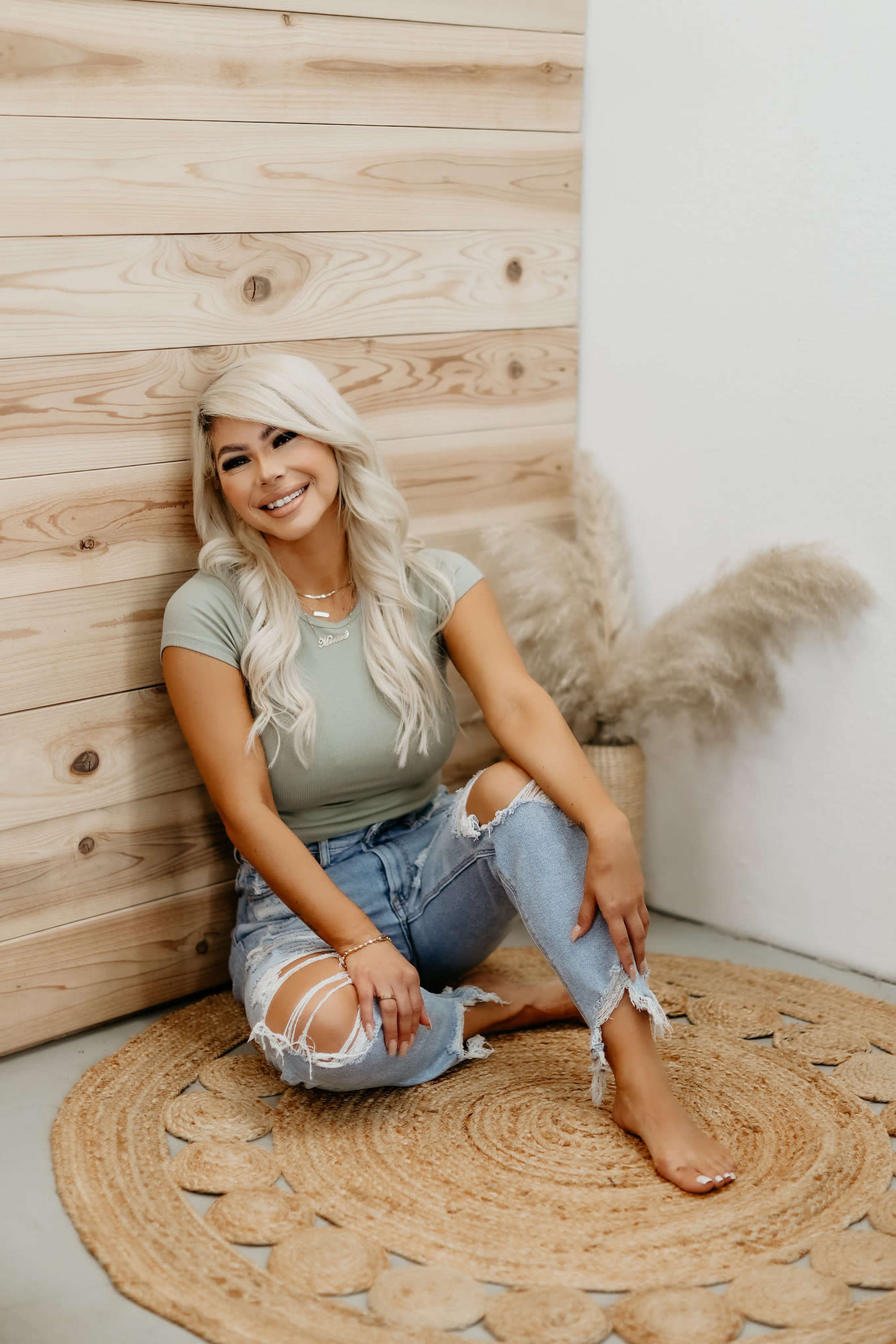 A woman with long, light-colored hair sits on a woven rug against a wooden wall, wearing a light green top and distressed jeans.