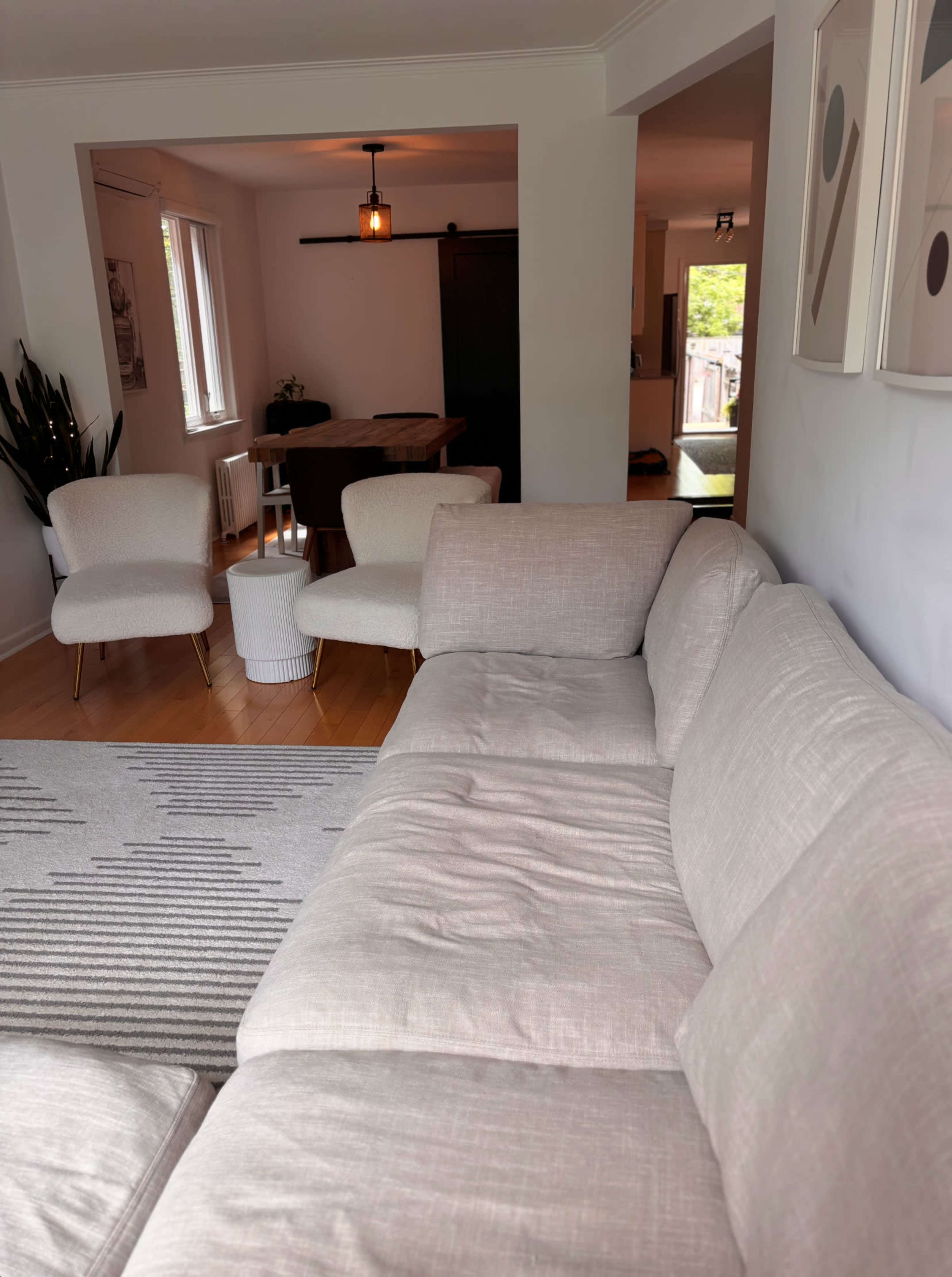 The image shows a modern living room with a light-colored sectional sofa, two contemporary chairs, and a dining area visible in the background.