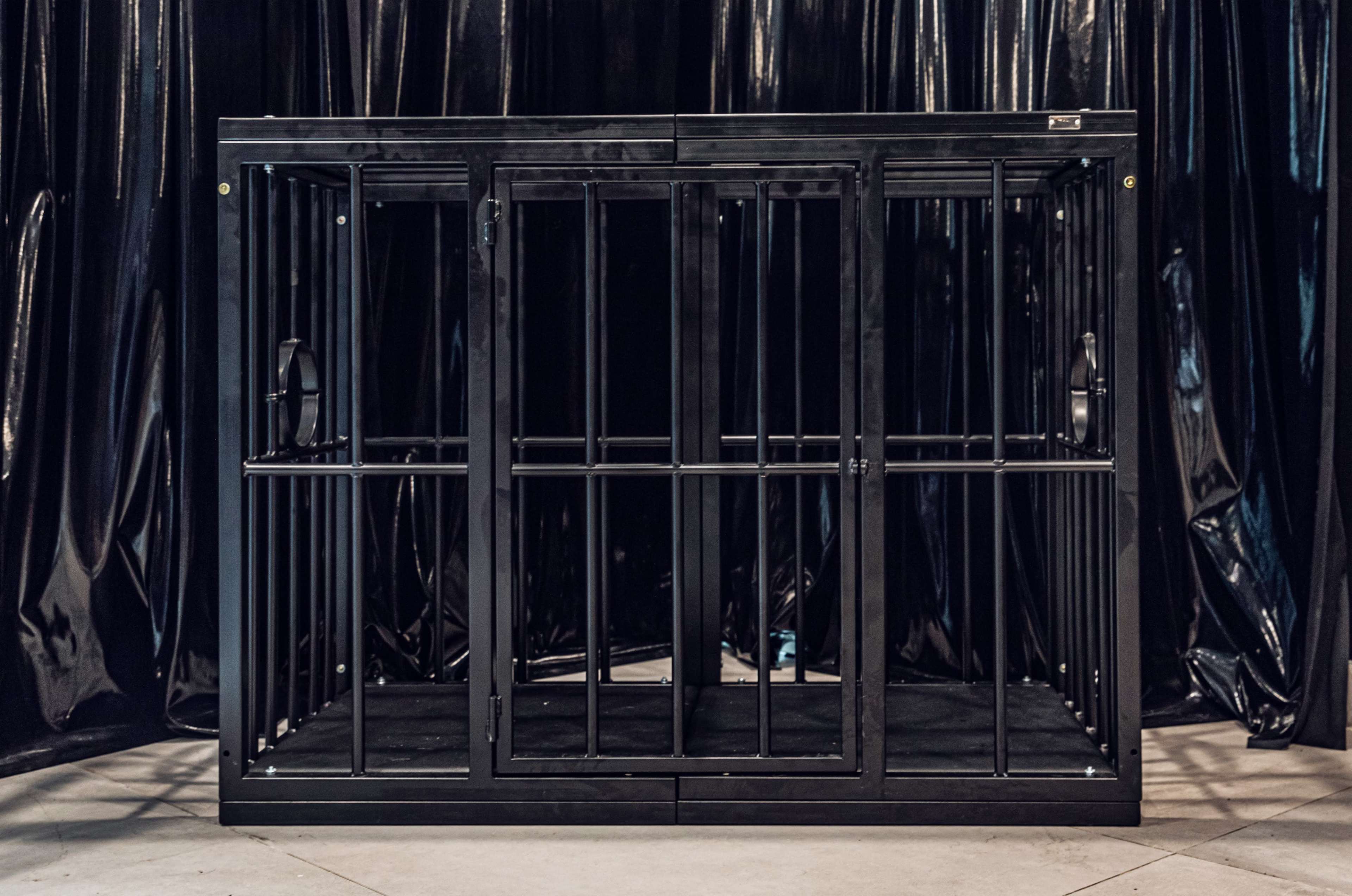 A large, empty black metal cage with barred doors, placed against a dark backdrop.