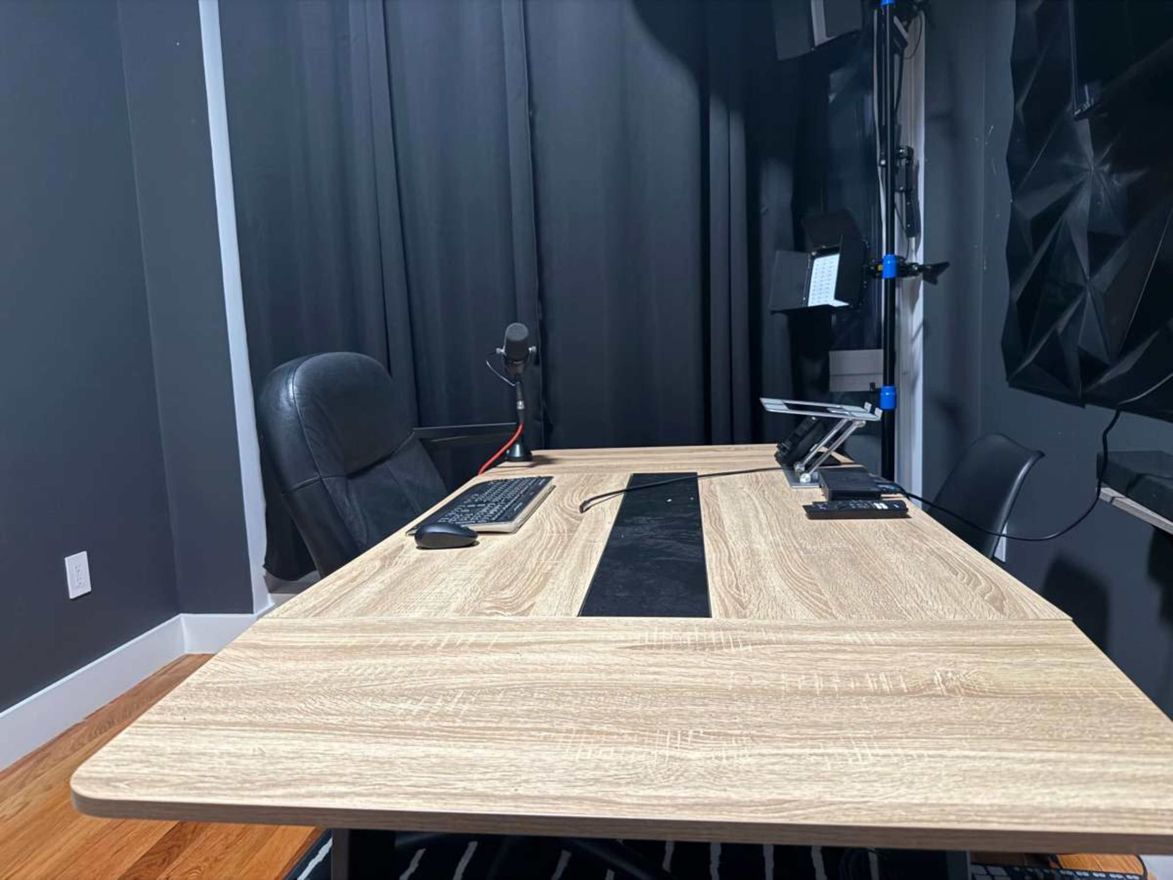 The image shows a modern office setup featuring a wooden desk with a microphone, a keyboard, and a chair, all positioned in front of a dark curtain.