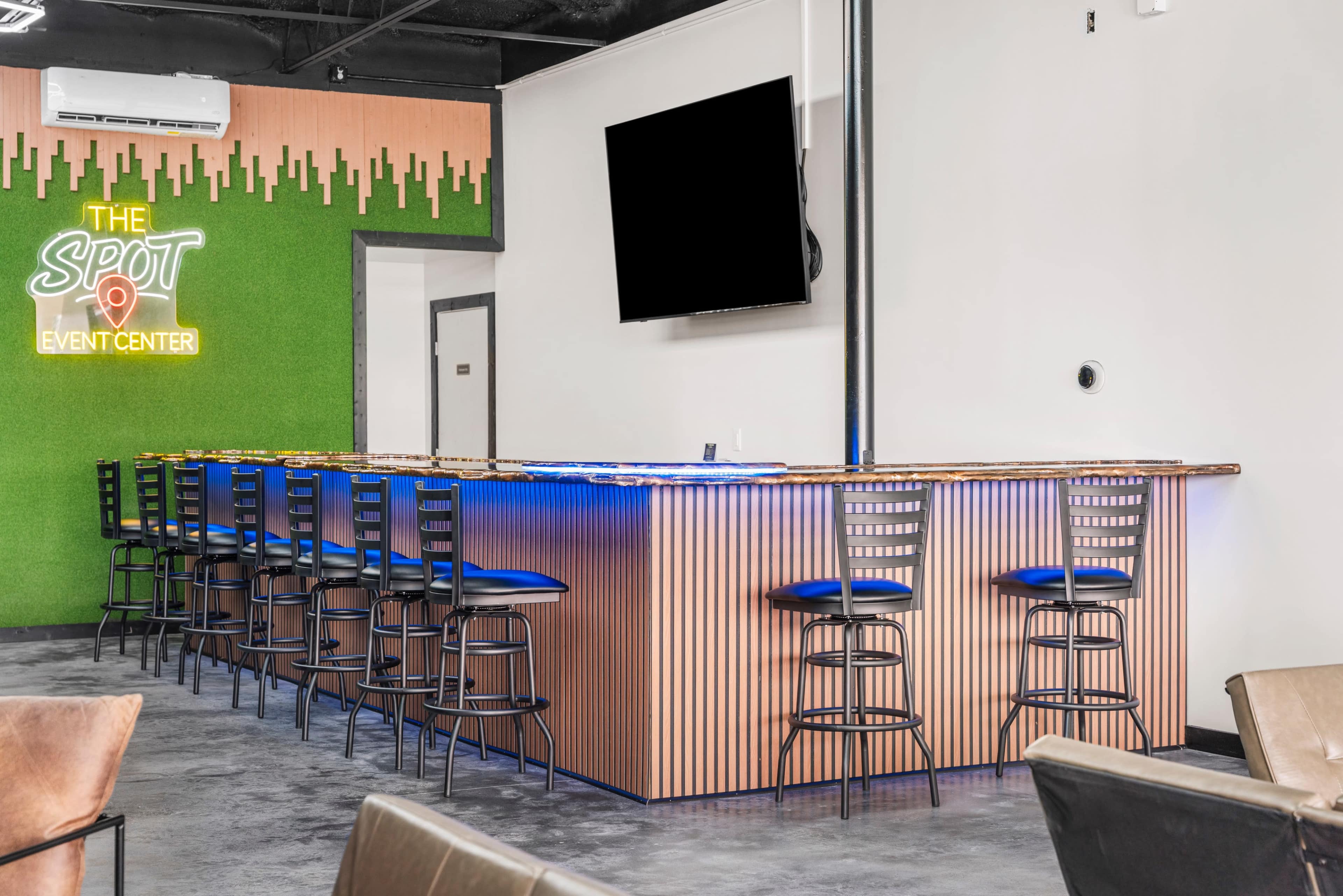 The image shows a modern bar area with a long wooden counter, high stools, and a large empty television screen on the wall.