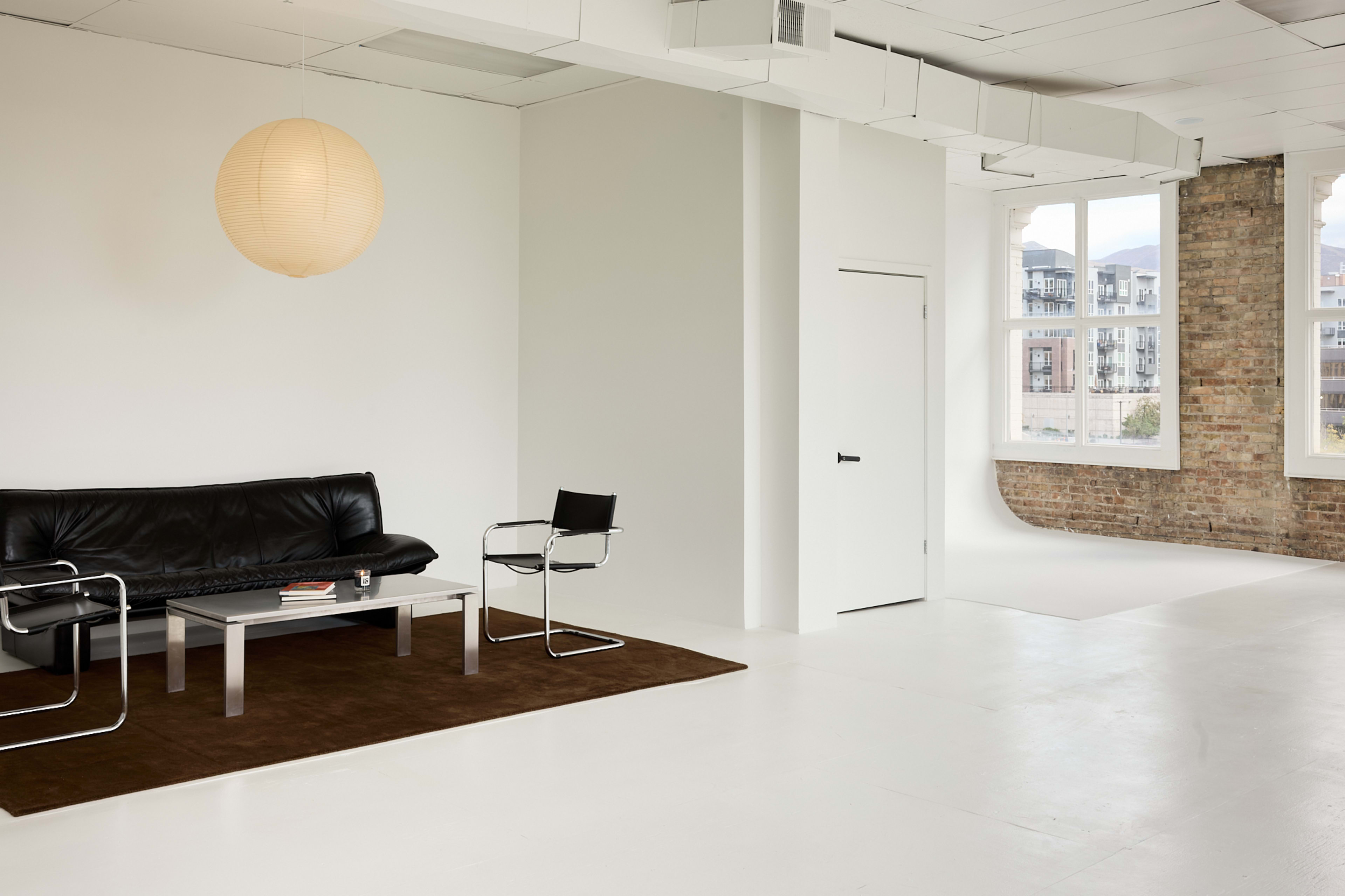 The image shows a modern, minimalist living space featuring a black leather sofa, a glass coffee table, and a round paper lantern hanging from the ceiling.