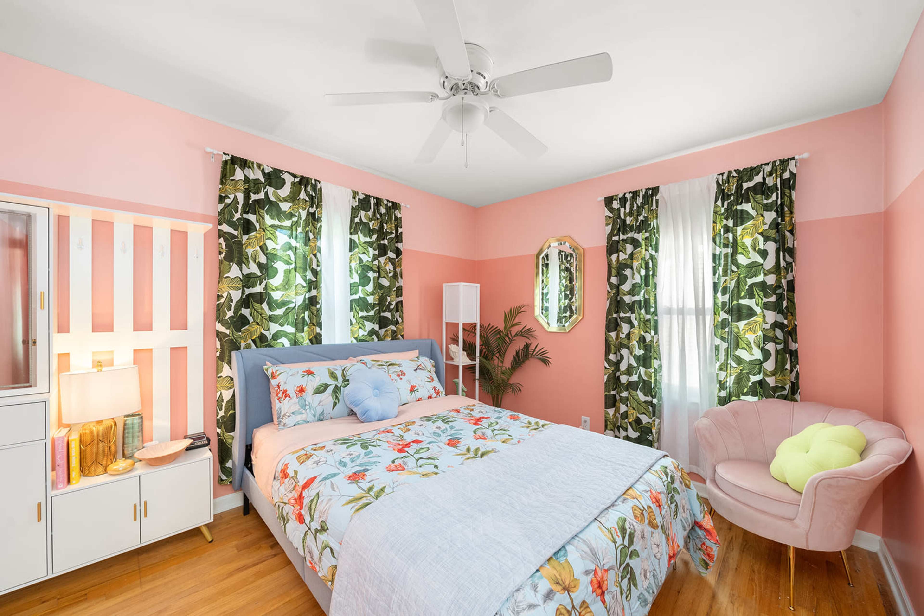 The bedroom features a bed with floral bedding, a pink accent wall, and large green plant-patterned curtains.