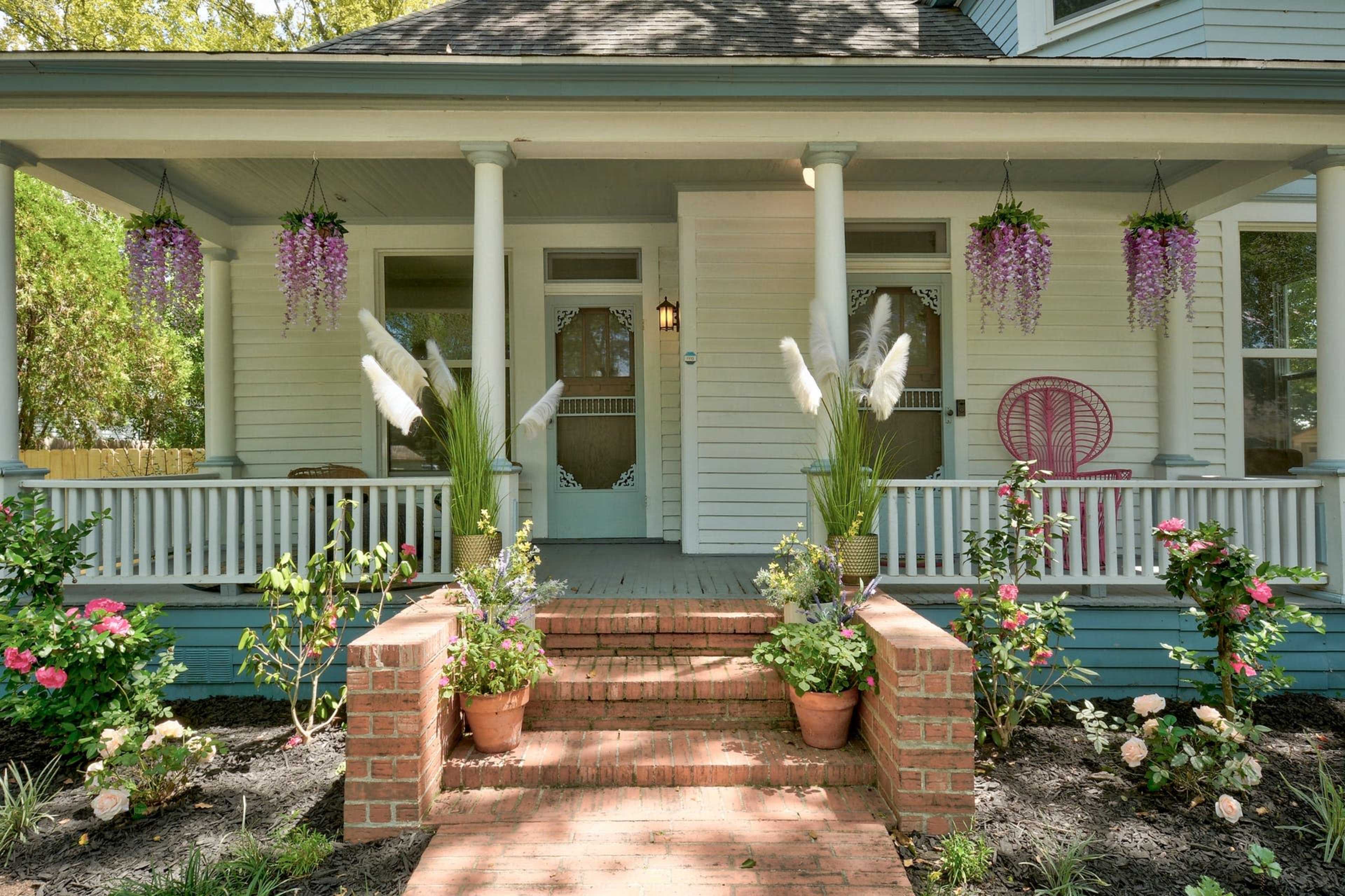 A porch of a light blue house adorned with hanging purple flowers, surrounded by potted plants and a brick pathway leading to the front door.