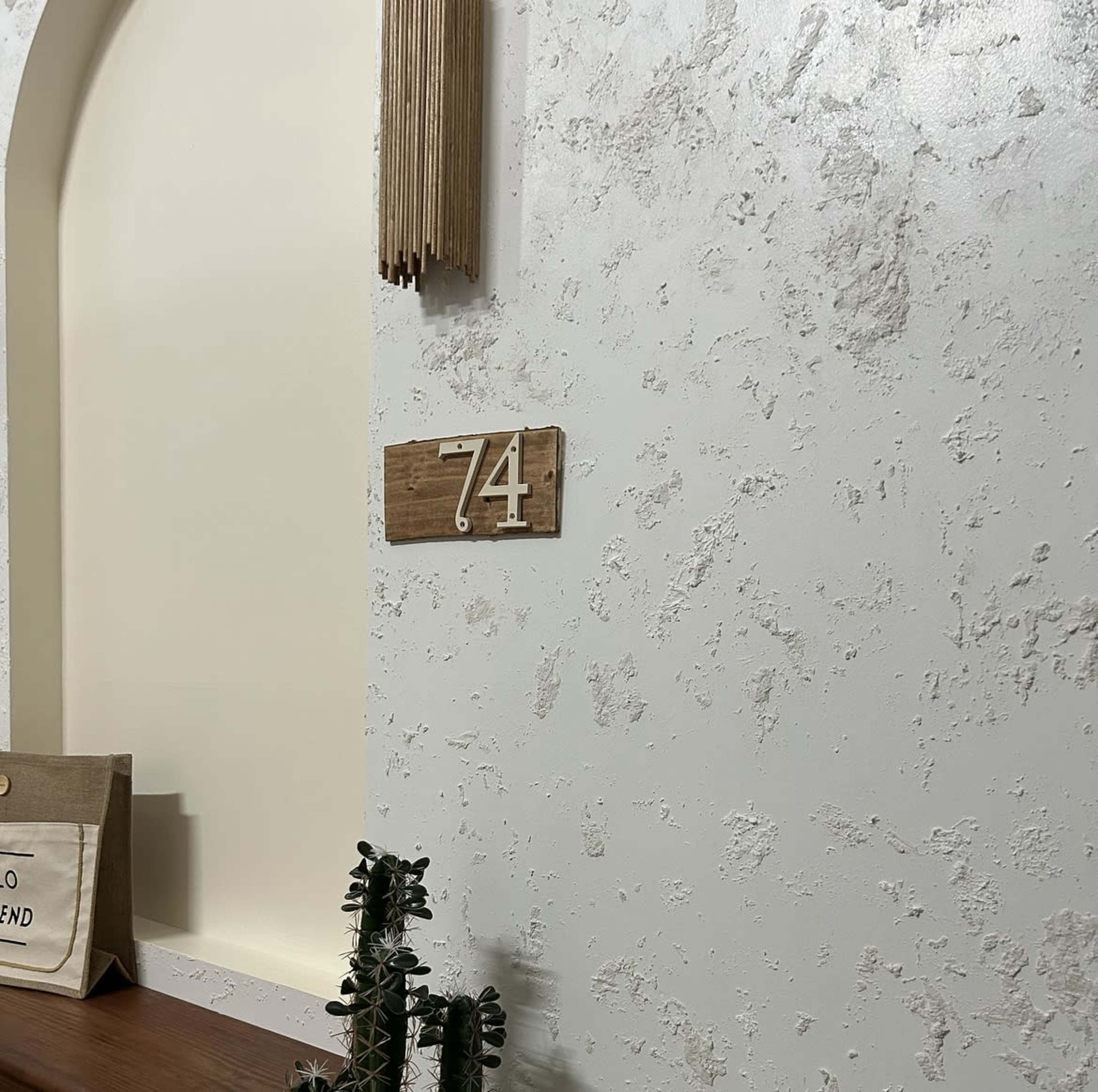 A wall with a textured finish, featuring the number "74" on a wooden plaque and a small potted cactus on a wooden shelf below.