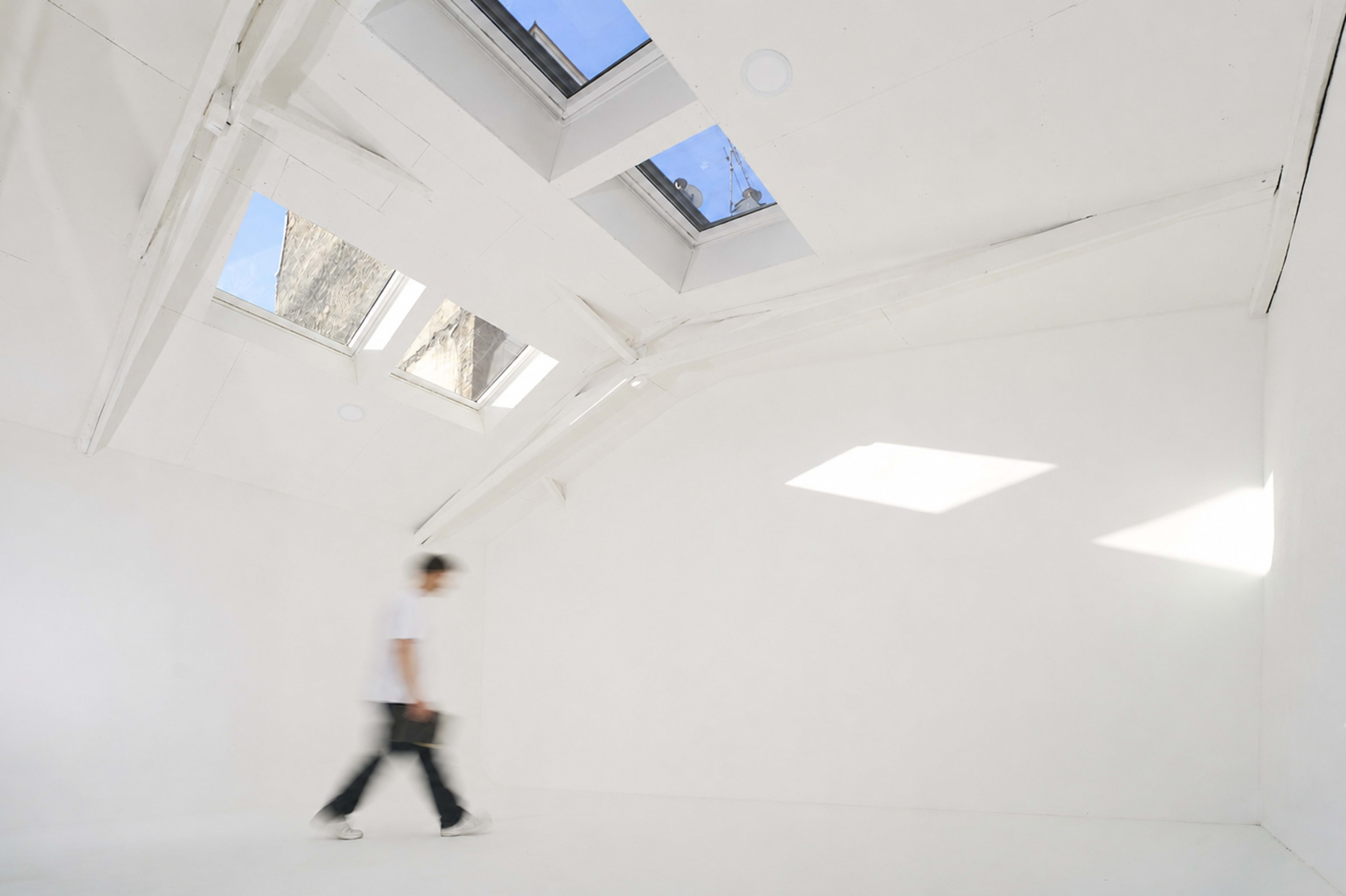 A person walks through a bright, minimally furnished room with white walls and skylights.