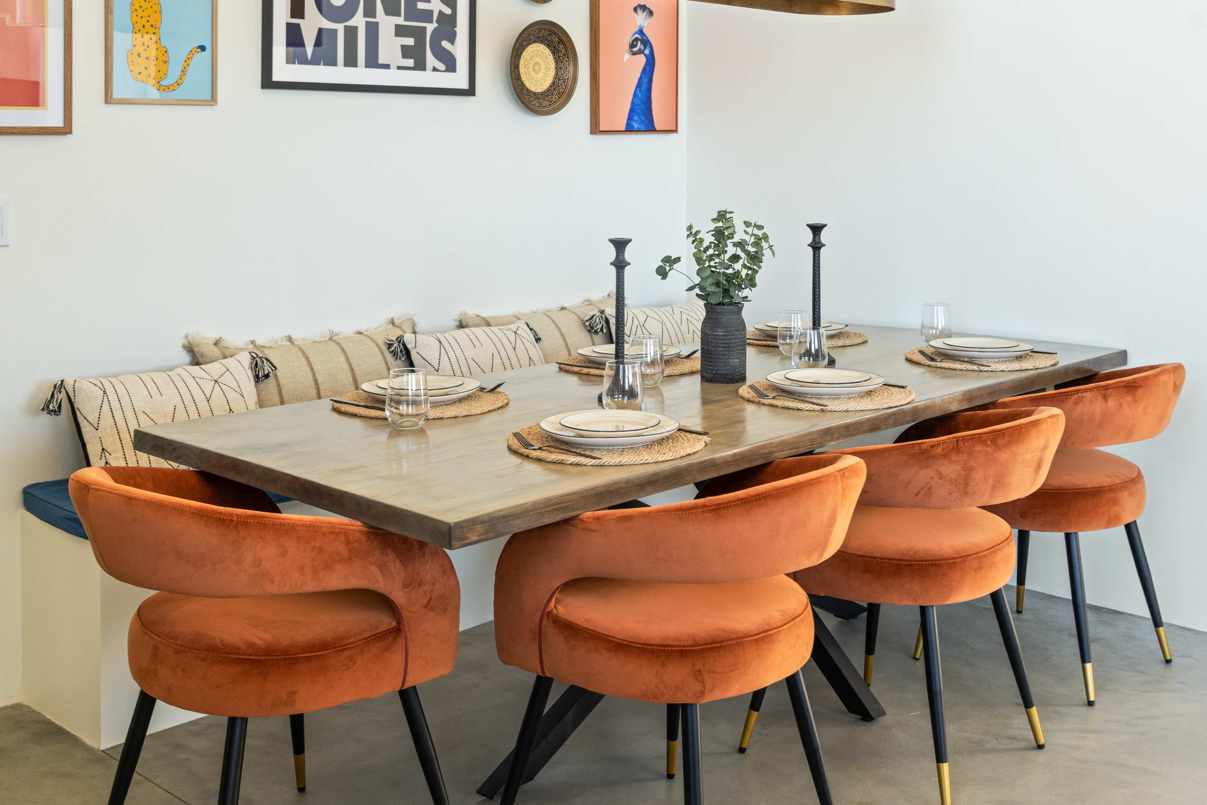 A long dining table with eight orange velvet chairs is set for a meal, featuring plates and glasses, alongside a decorative wall with framed art.