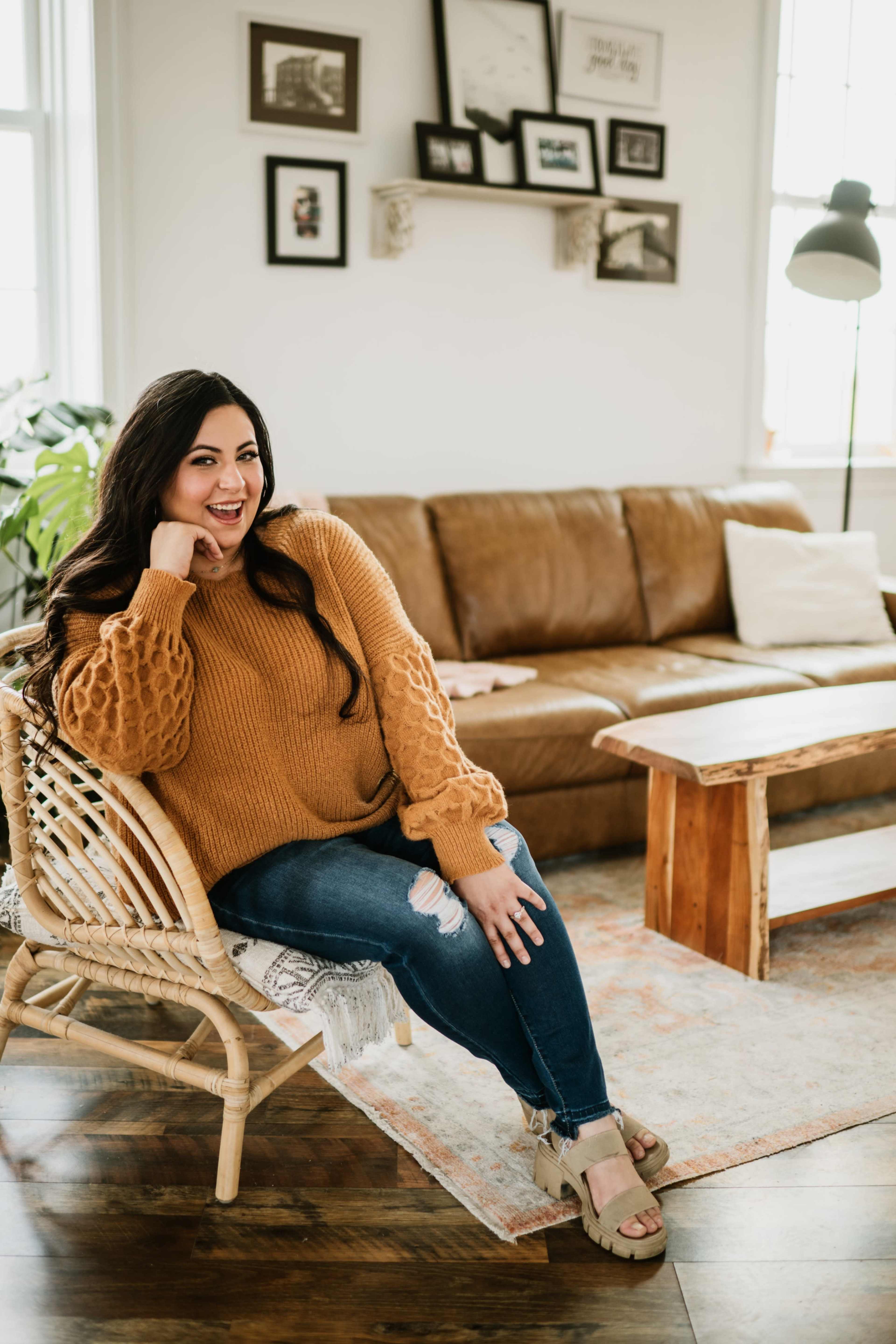 A woman sits in a woven chair, wearing a cozy sweater and jeans, in a warmly decorated living room with a brown sofa and wooden coffee table.