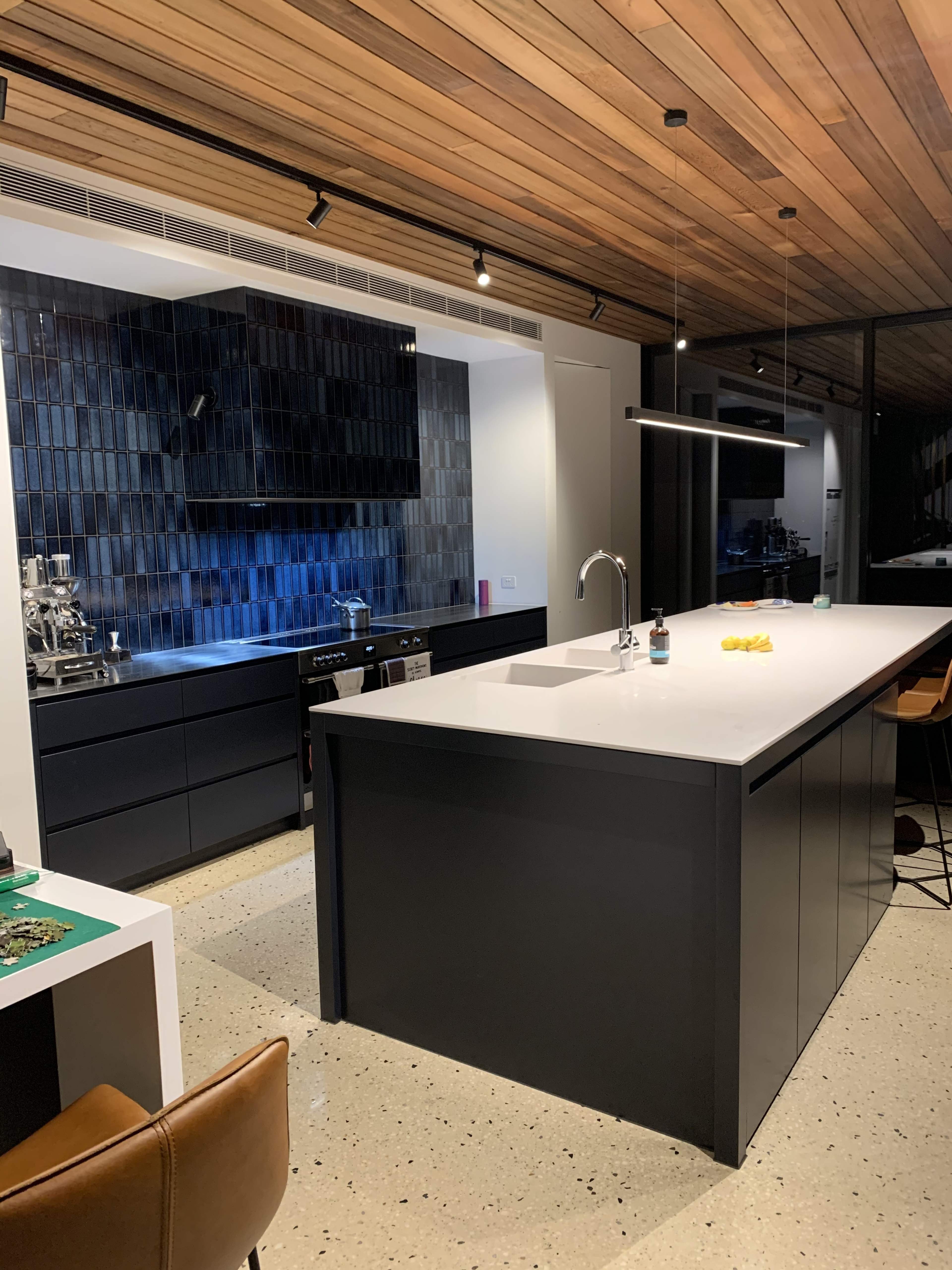 A modern kitchen featuring a central island, black cabinetry, and a wooden ceiling with recessed lighting.