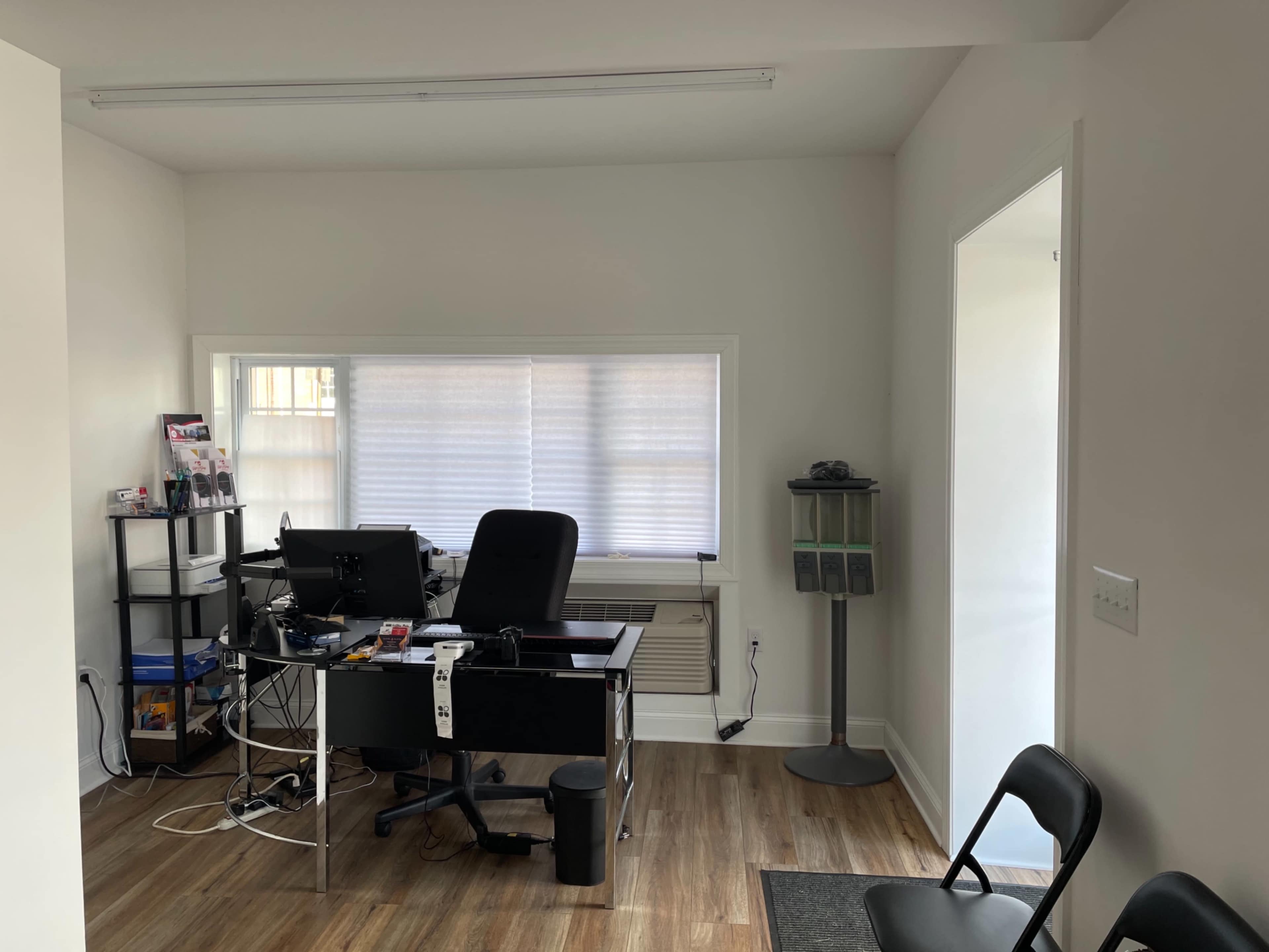 A minimalistic office space featuring a black desk with a computer, a chair, and a bookshelf, along with a small waiting area with two chairs.
