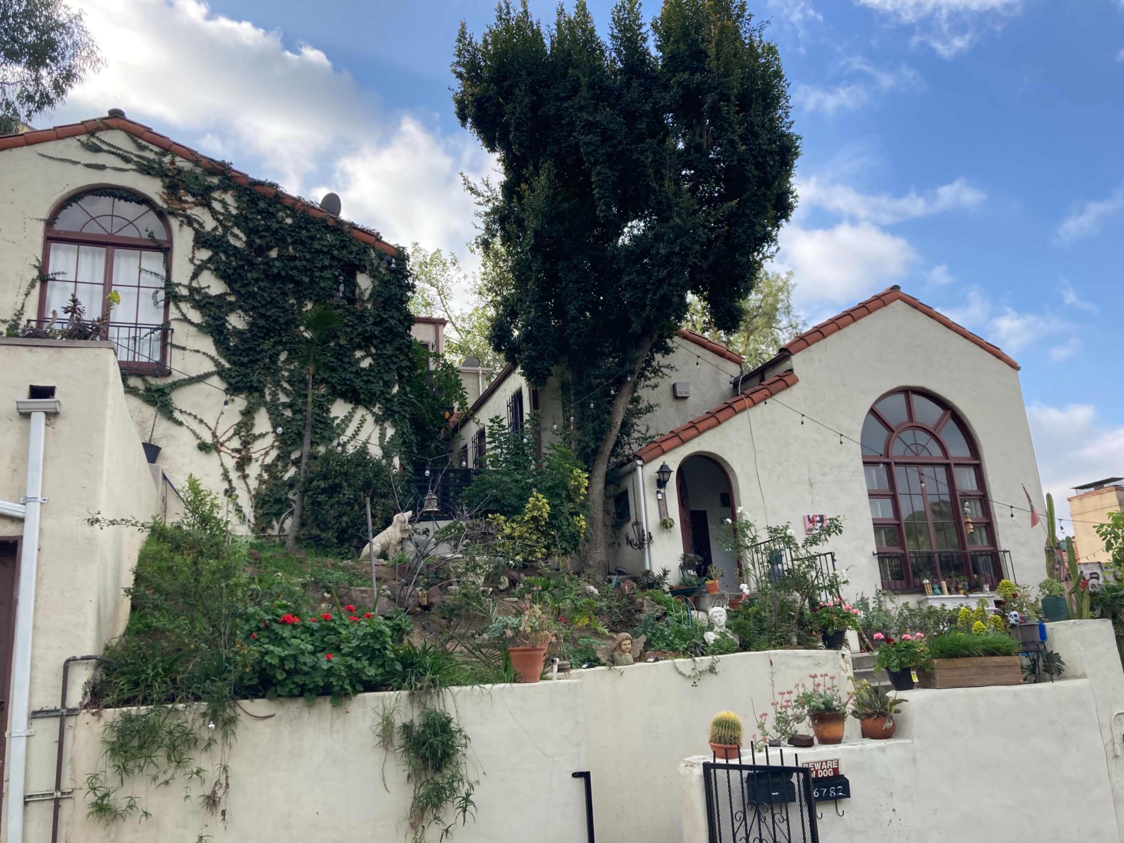 A hillside house with white walls, red-tiled roofs, and various plants in pots surrounding the entrance.