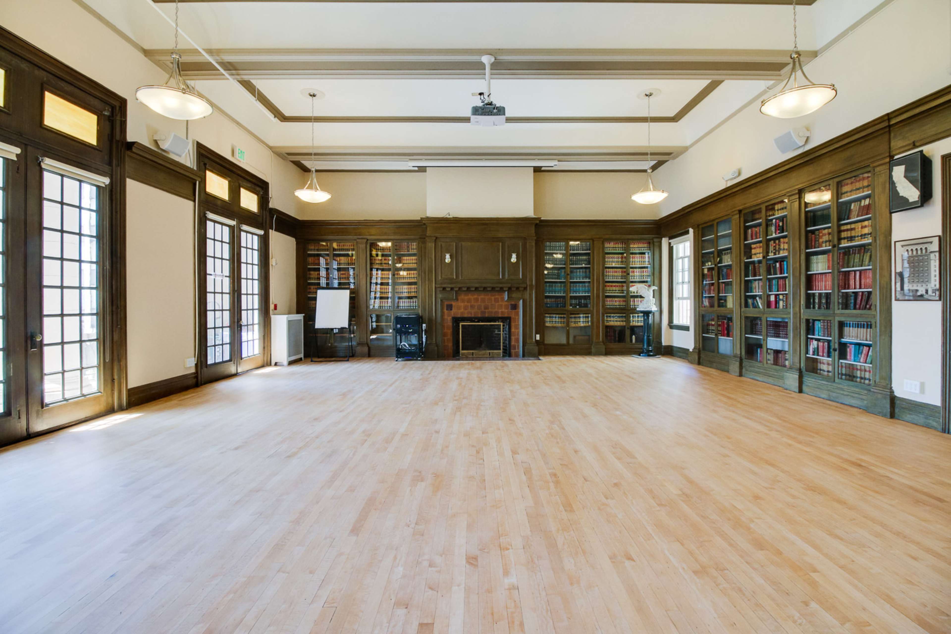 A large, empty room features wooden floors, bookshelves lining the walls, and multiple doors leading outside.