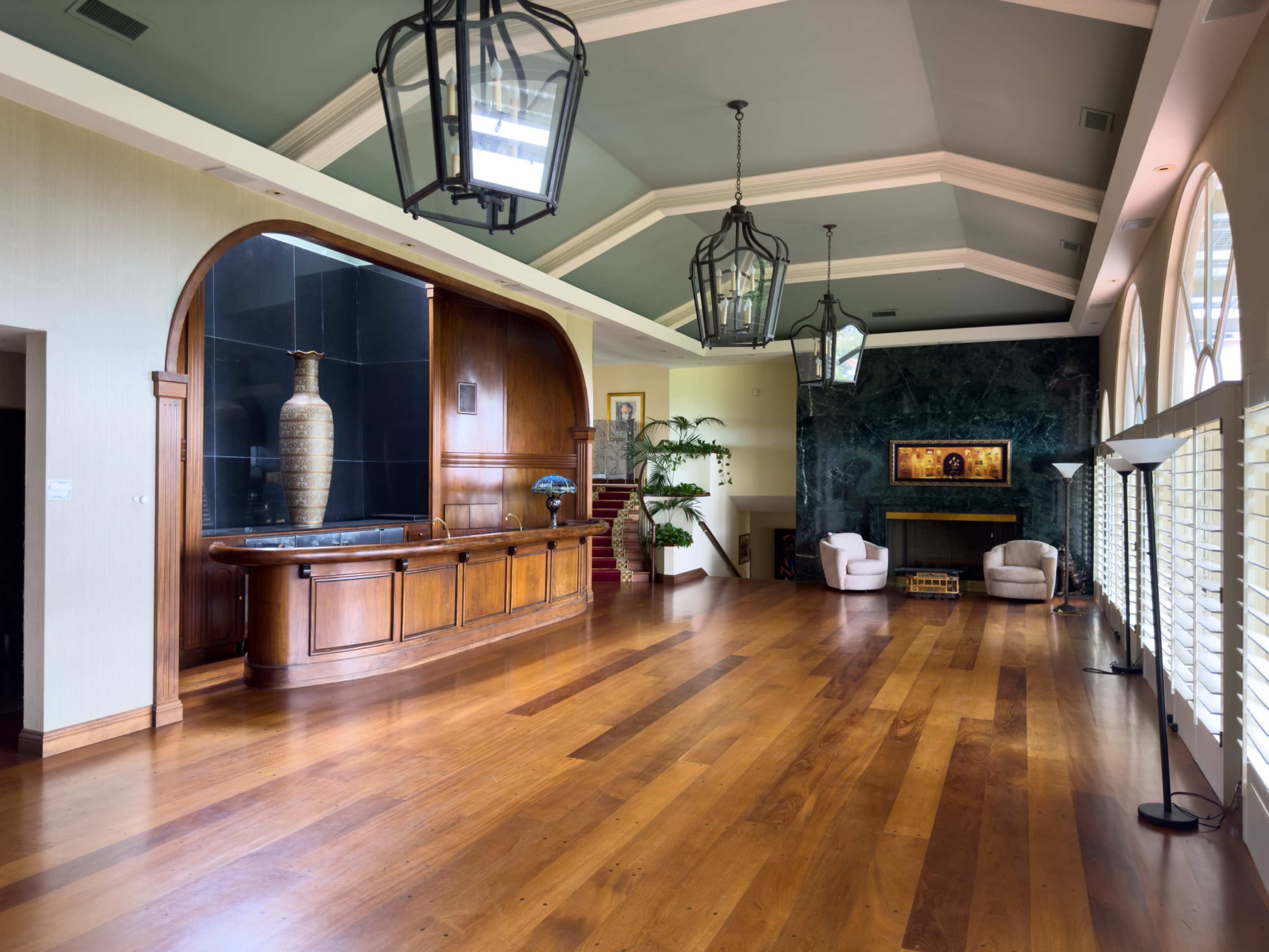 The image shows a spacious living area featuring a wooden floor, large arched windows, and a high ceiling with pendant lights, along with a bar area and modern furnishings.