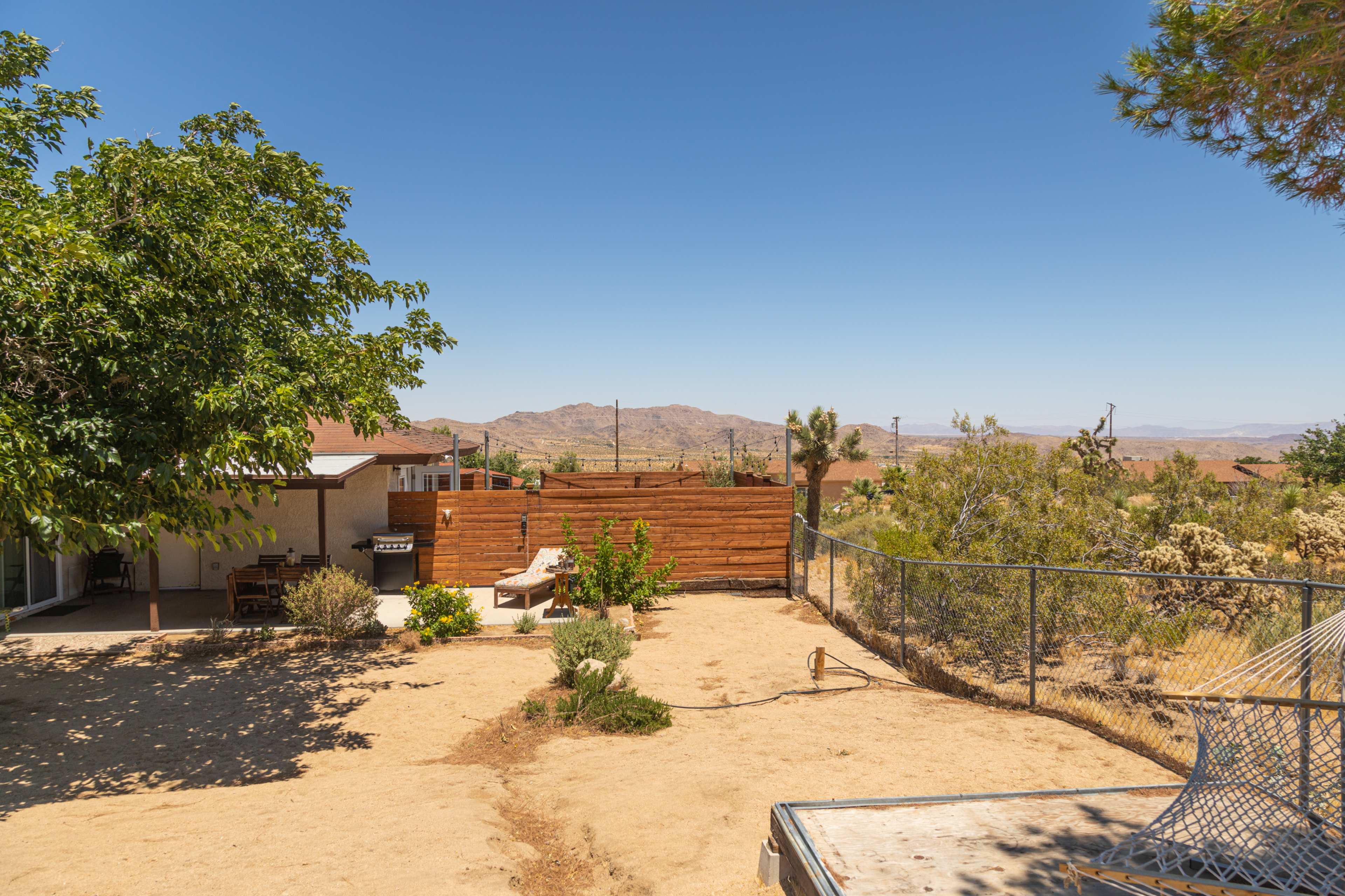 The scene shows a desert landscape with a fenced backyard, featuring a few plants and wooden fencing, and mountains in the background under a clear blue sky.