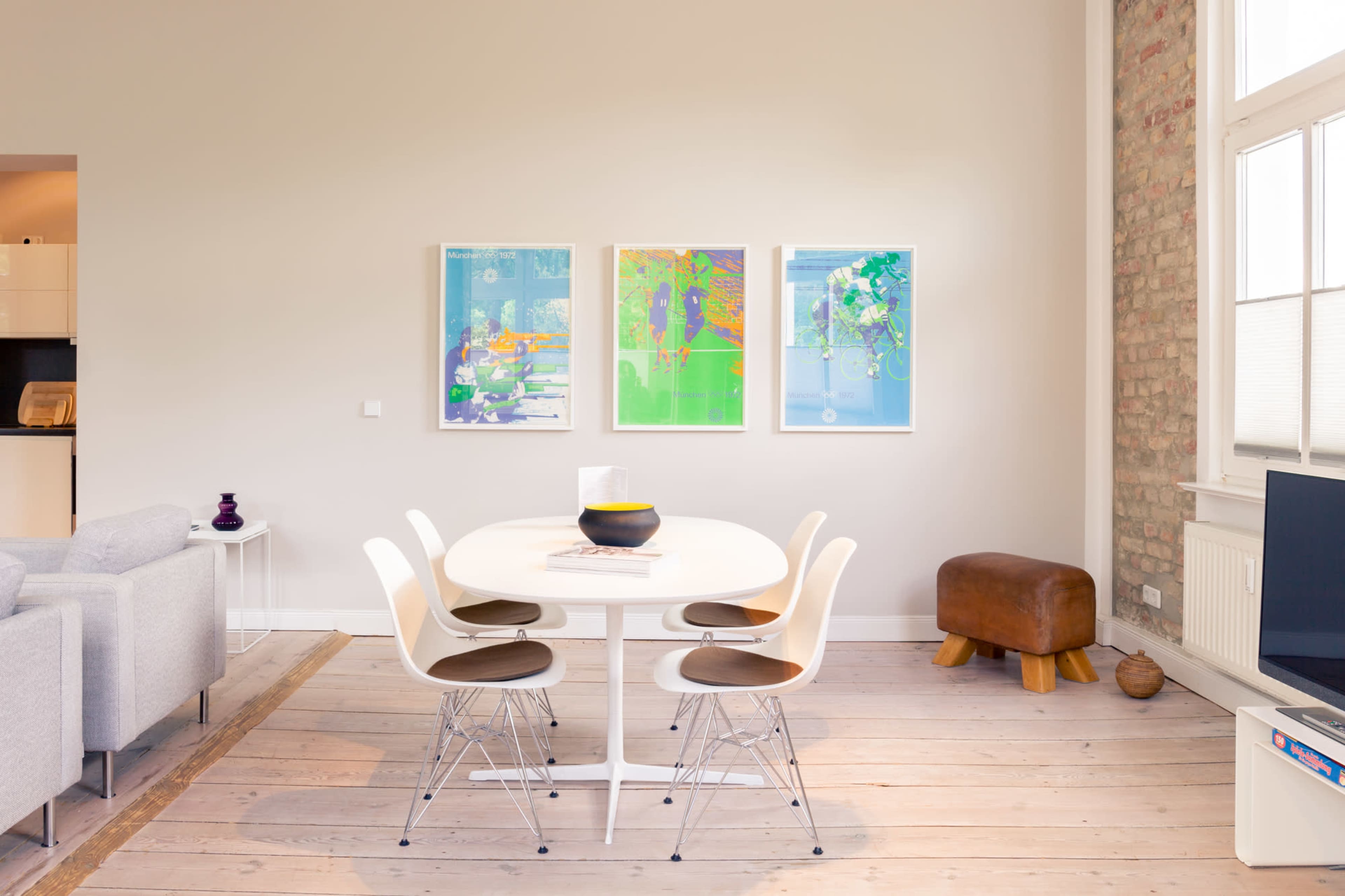 A modern dining area features a round white table surrounded by four chairs, with three colorful artworks on the wall and a wooden stool nearby.
