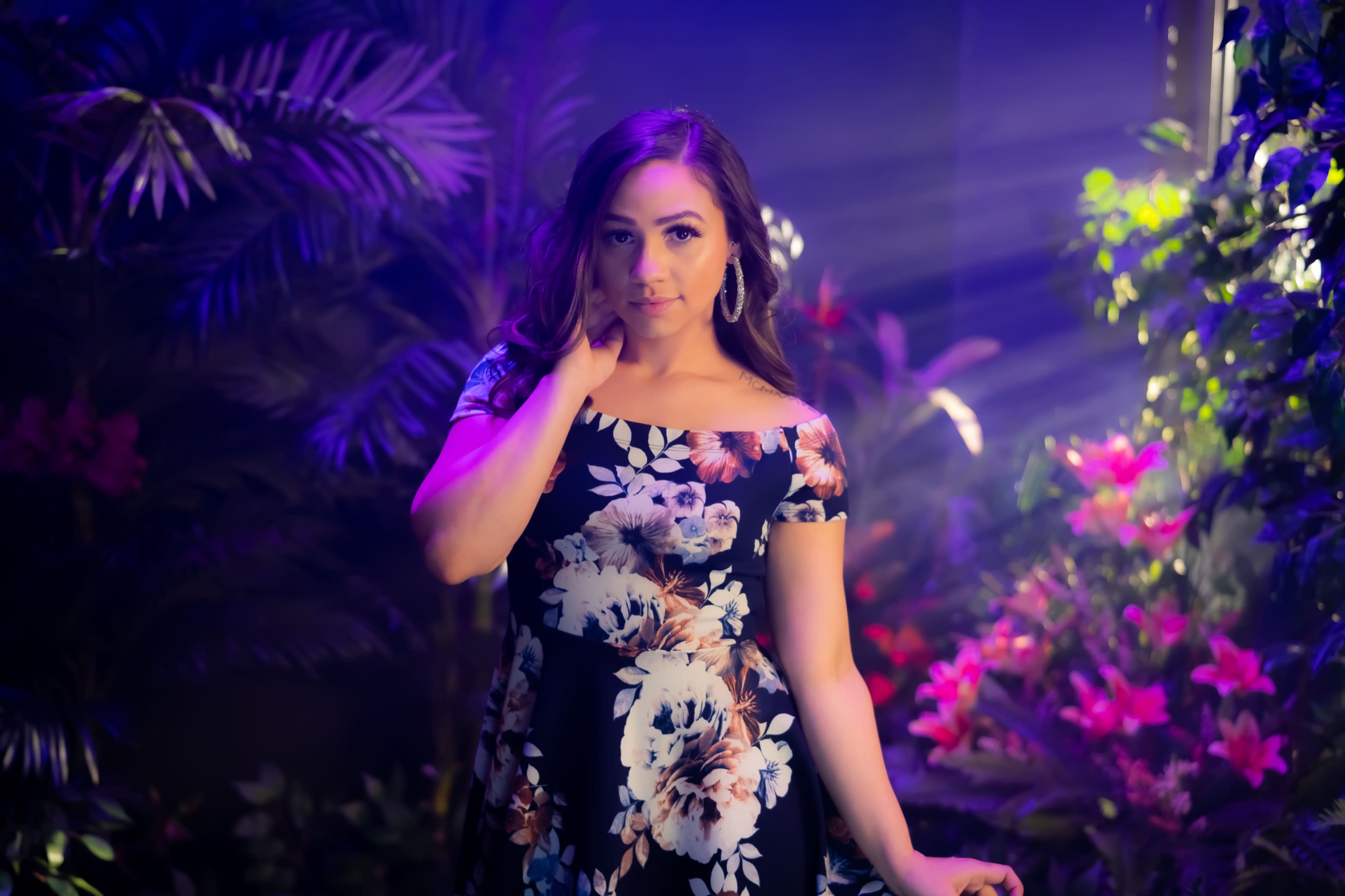 A woman in a floral dress poses amidst vibrant plants and soft, colorful lighting.