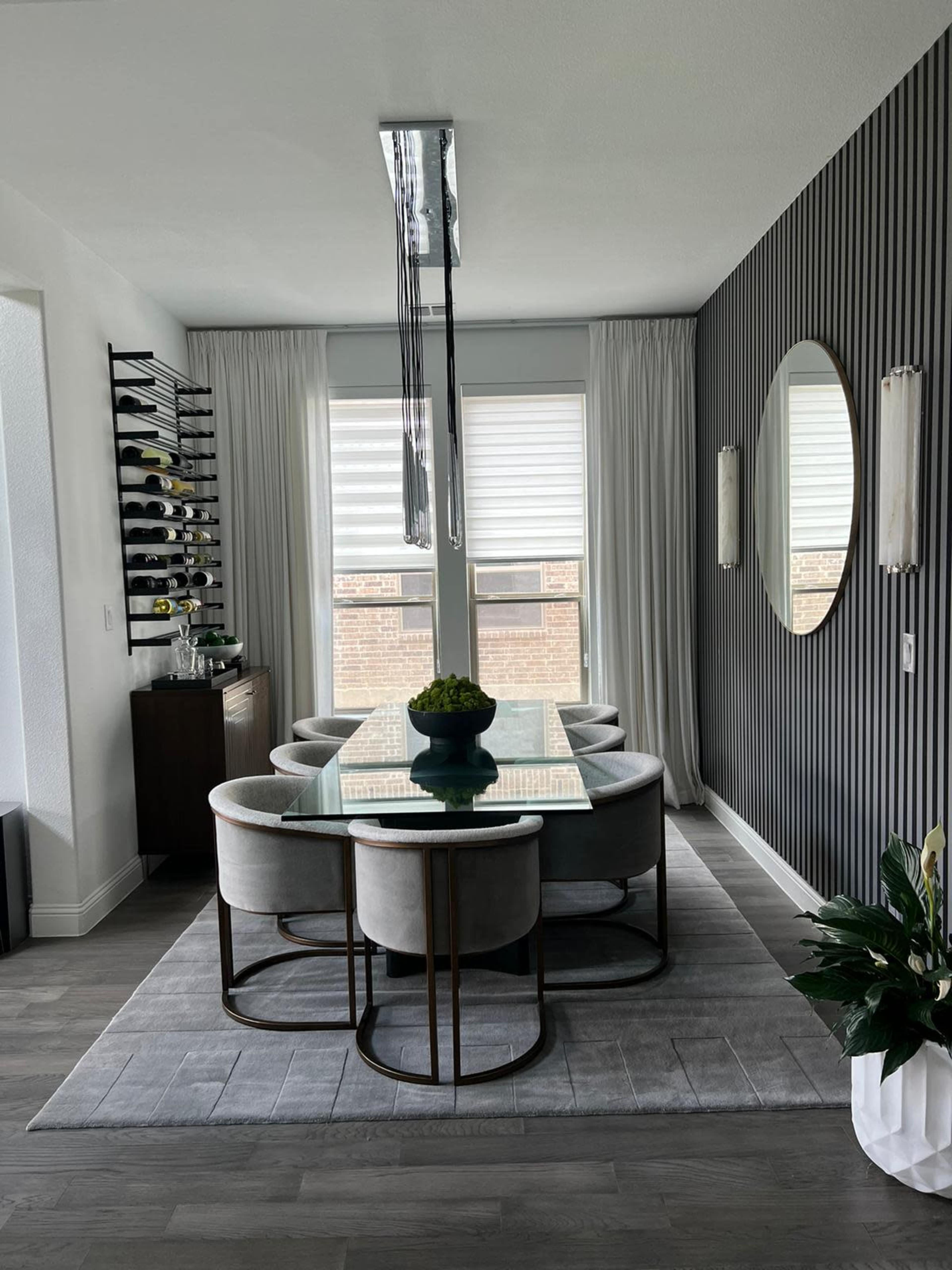 A modern dining room features a glass table surrounded by circular gray chairs, with large windows covered by sheer curtains and a decorative wall mirror.