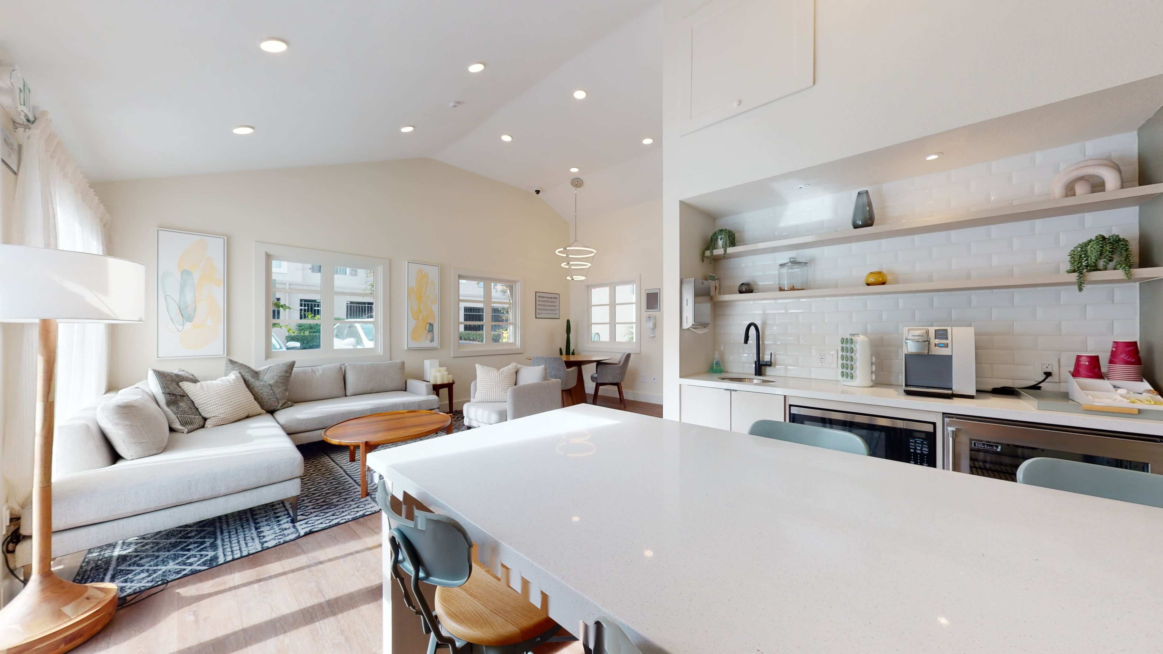 A modern living space features a light-filled open layout with a seating area, a kitchen island, and shelving displaying decorative items.