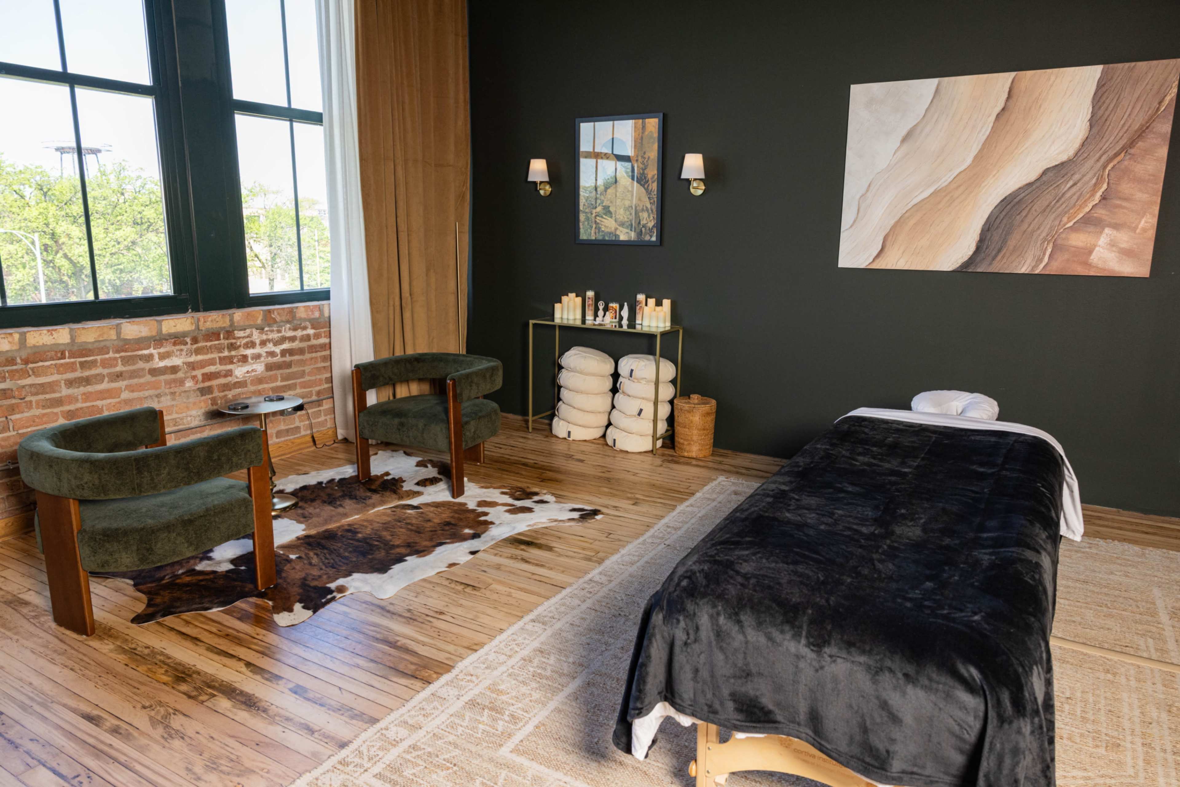 The image features a massage room with a massage table, two green armchairs, a cowhide rug, and shelves holding towels and skincare products.