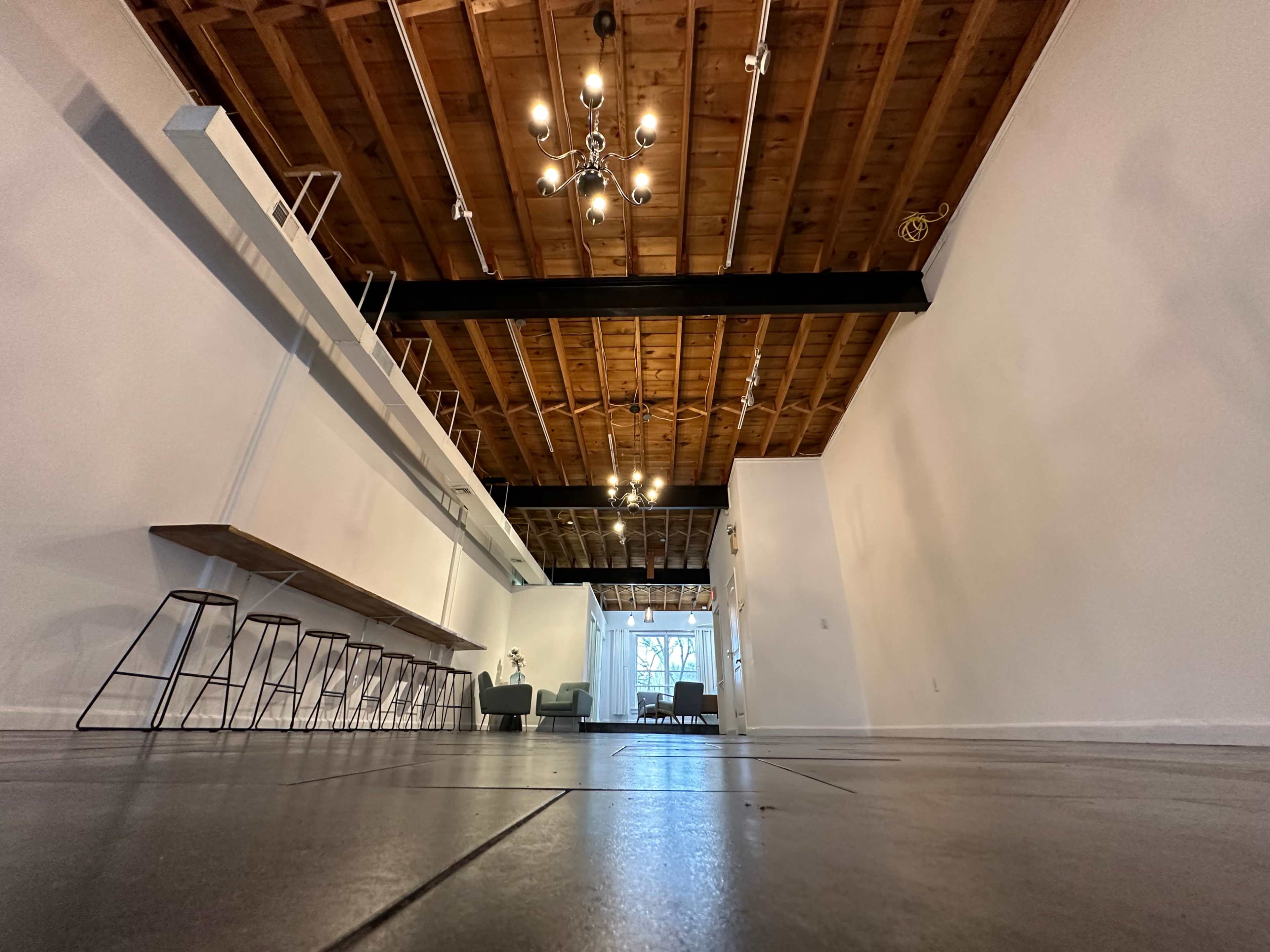 A spacious interior with exposed wooden beams, a long bar area with stools, and a wide entrance leading to a well-lit area.