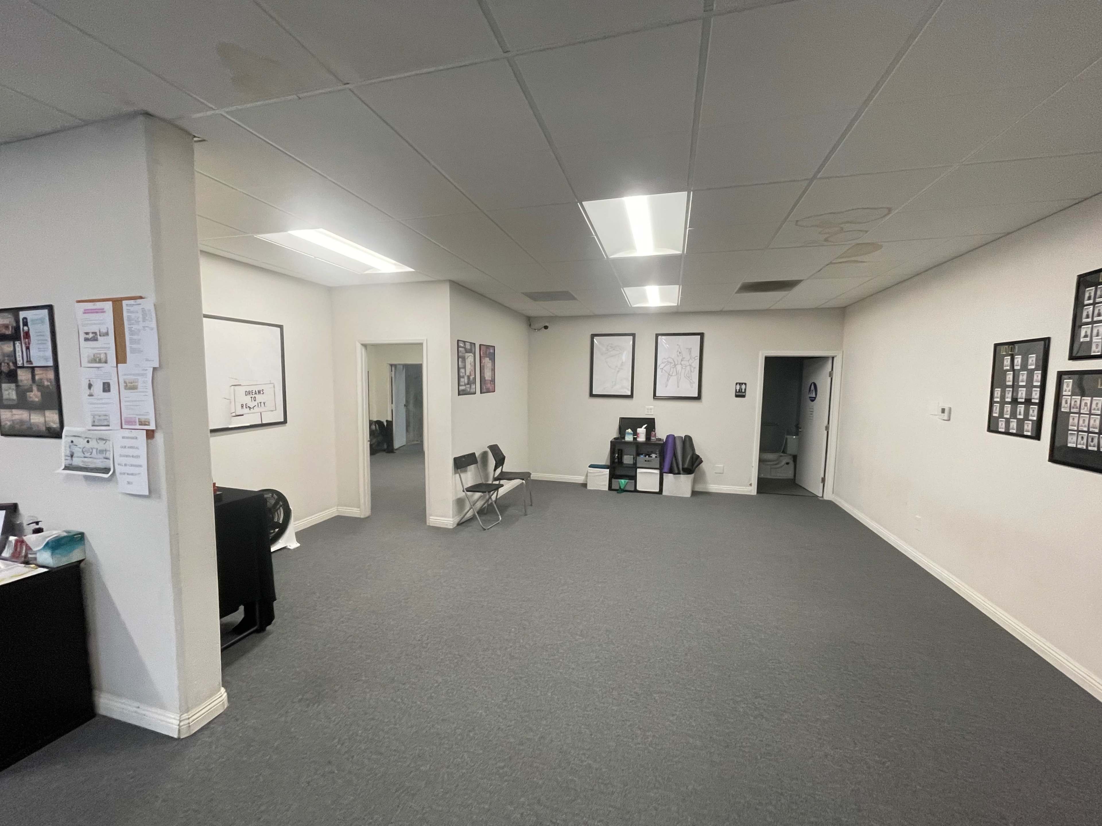 An empty office space features gray carpeting, white walls, fluorescent lighting, and several framed pictures on the walls.