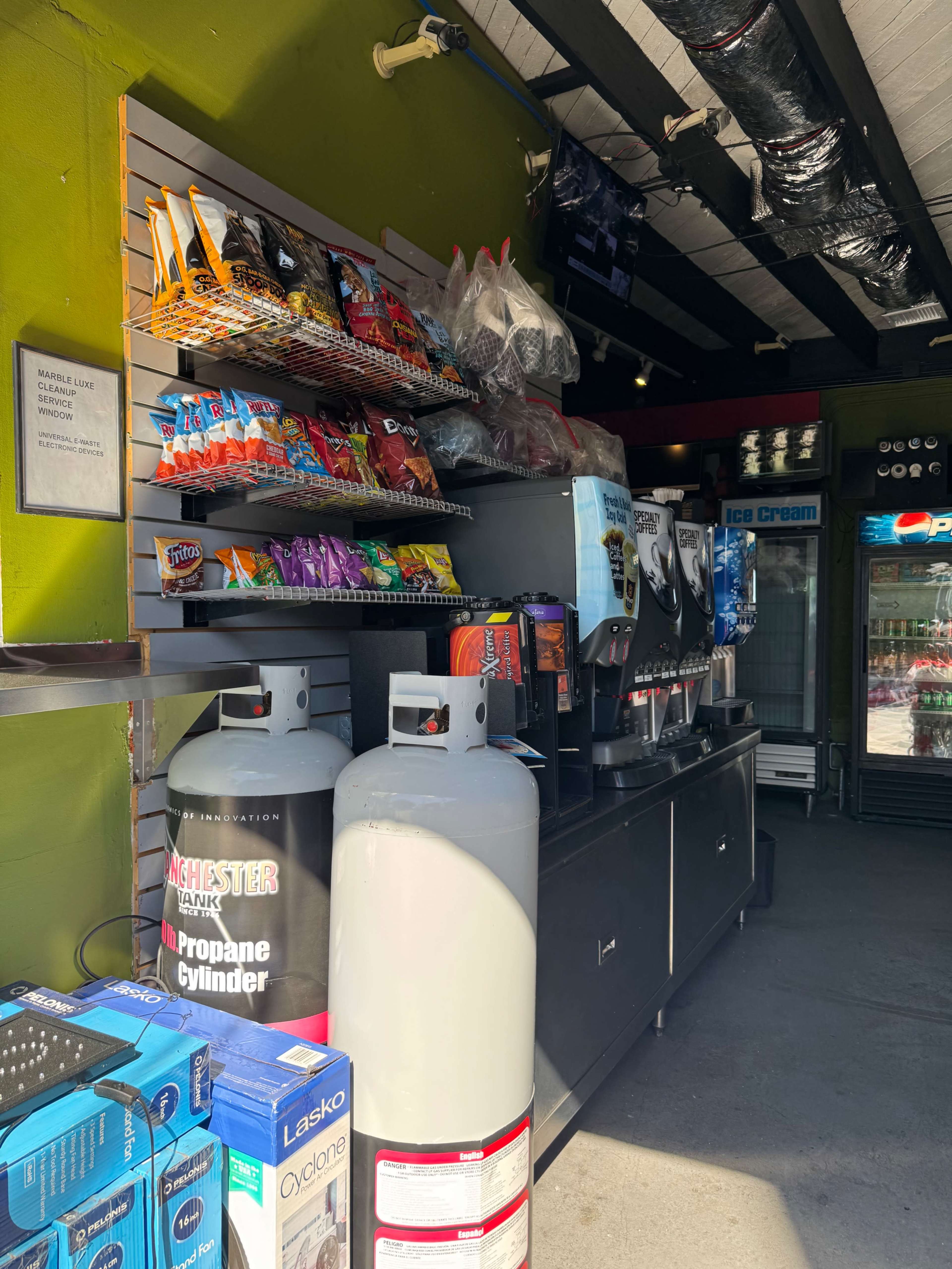 The image shows a convenience store counter displaying snacks, drinks, and propane tanks, with various items organized neatly on shelves.