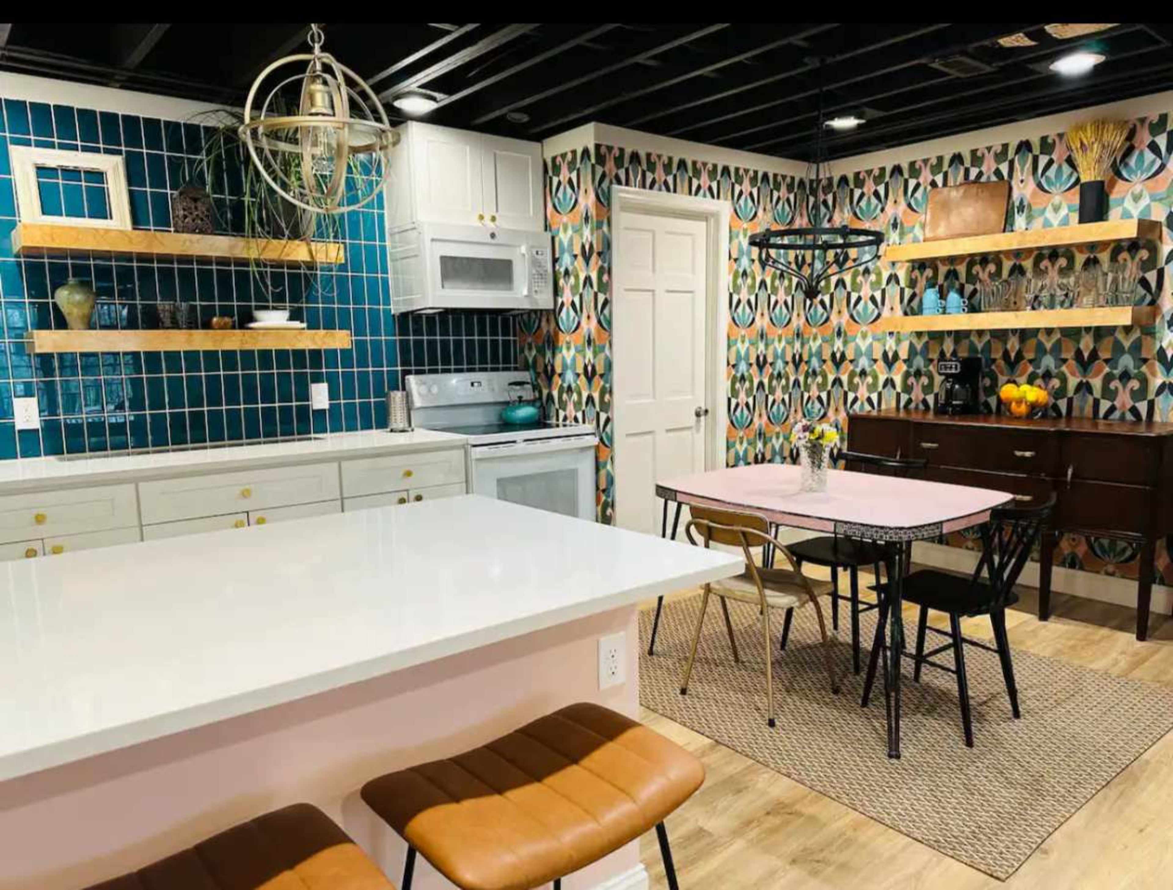 The image shows a modern kitchen with teal and patterned wallpaper, wooden shelves, and a dining table surrounded by black chairs.