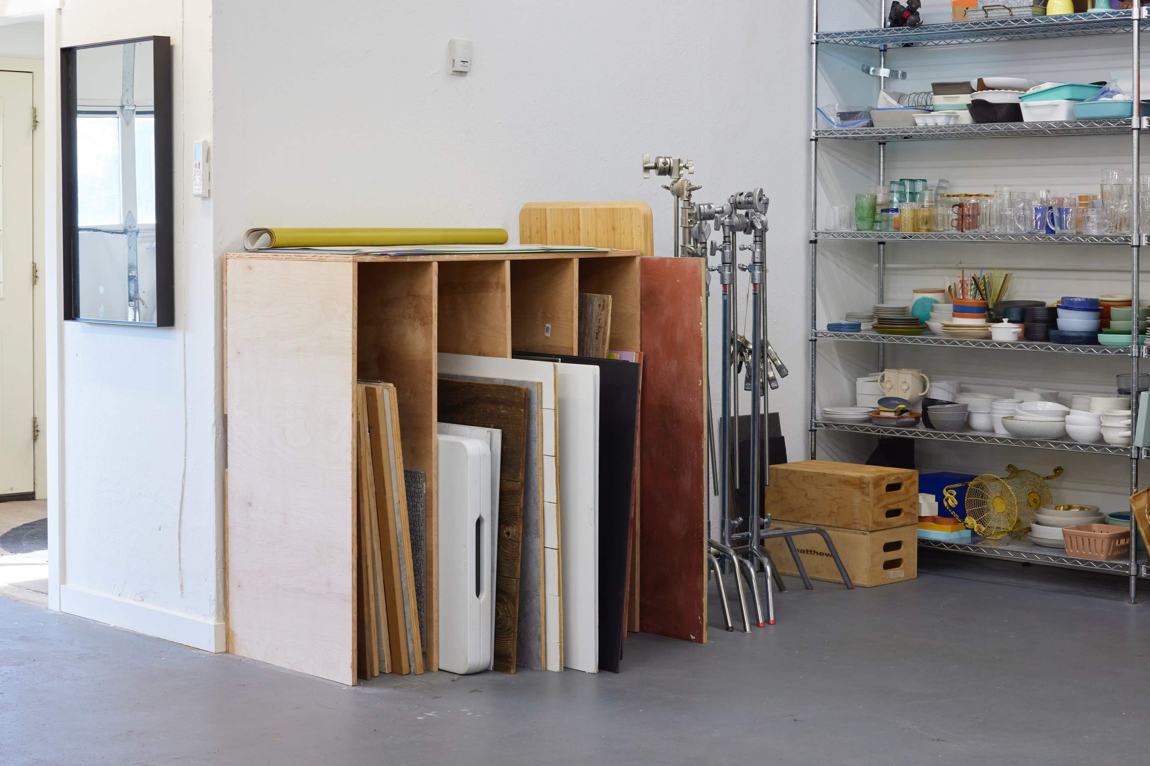 A collection of wooden boards is stacked next to a shelving unit holding various kitchen items and utensils in a well-lit room.