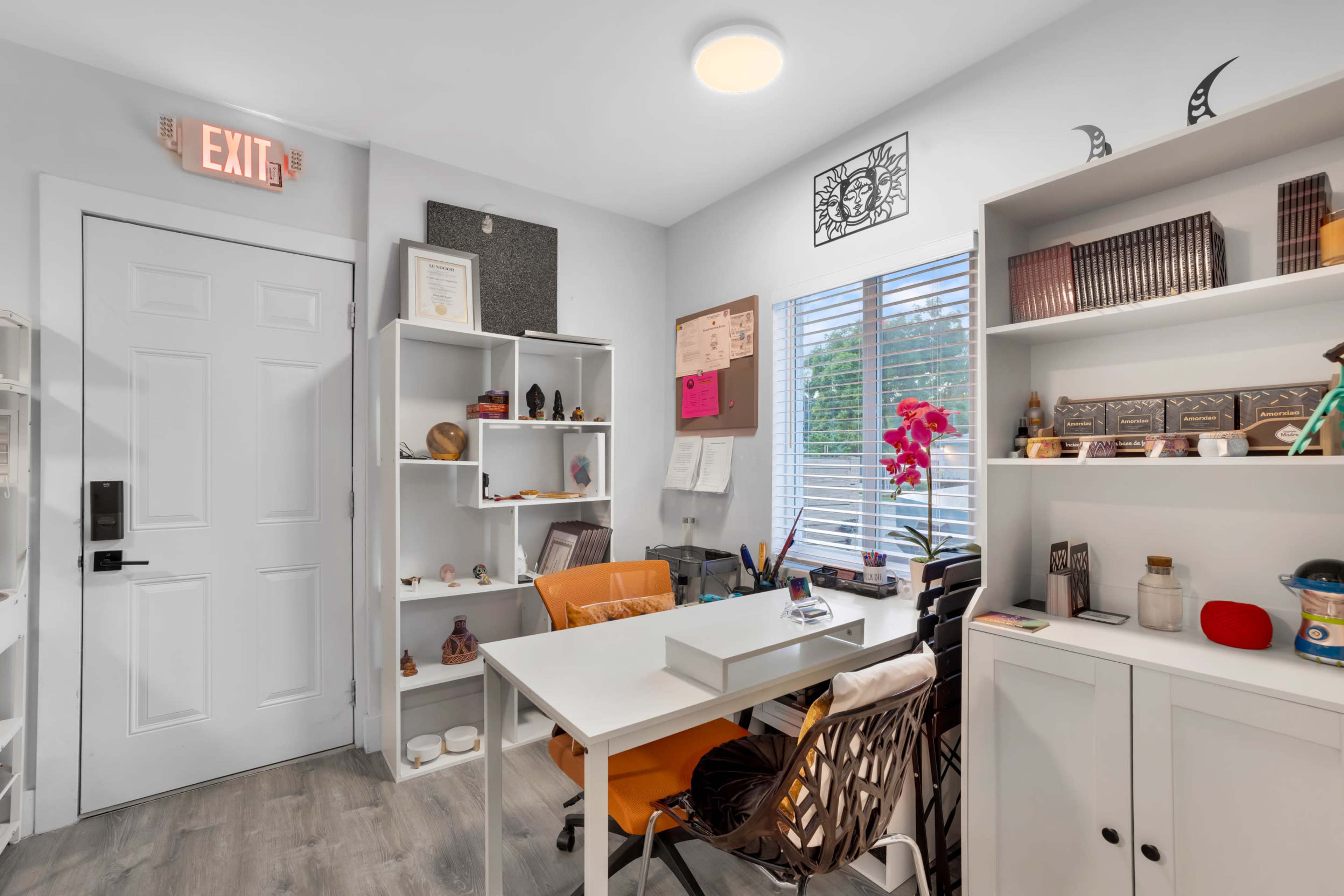 The image shows a small office space featuring a white desk and chair, a shelf with decorative items, and a window with natural light.