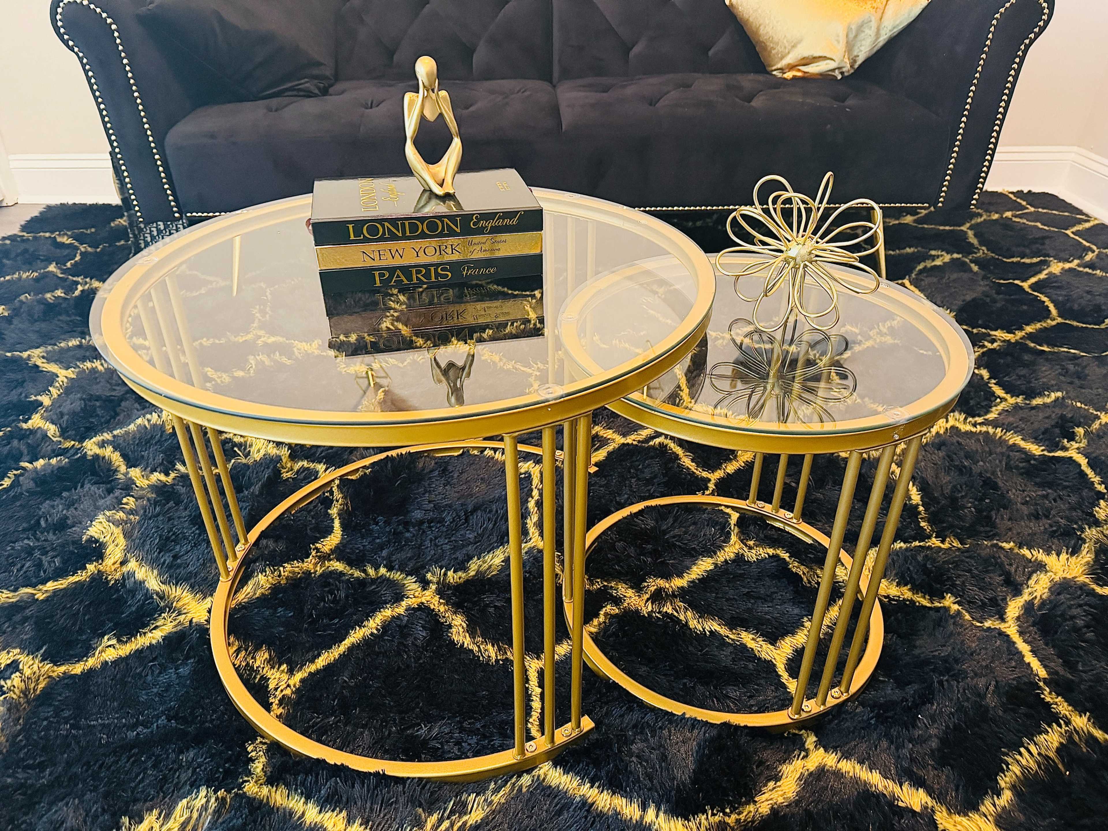 The image shows a pair of circular coffee tables with gold metal frames and glass tops, placed on a black and gold patterned rug in front of a dark sofa.