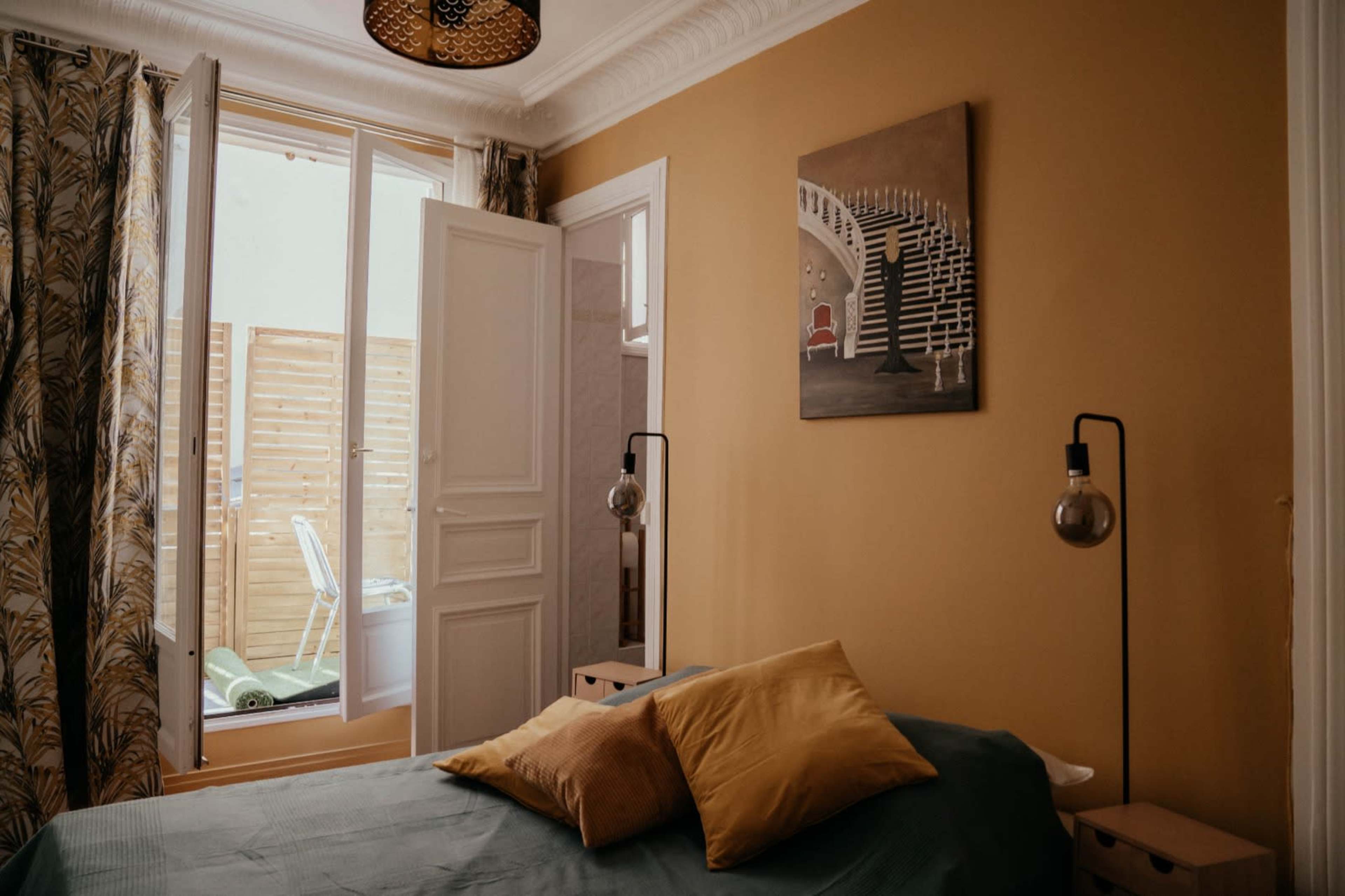 A cozy bedroom features a bed with yellow cushions, an artwork on the wall, and an open door leading to a small outdoor space.
