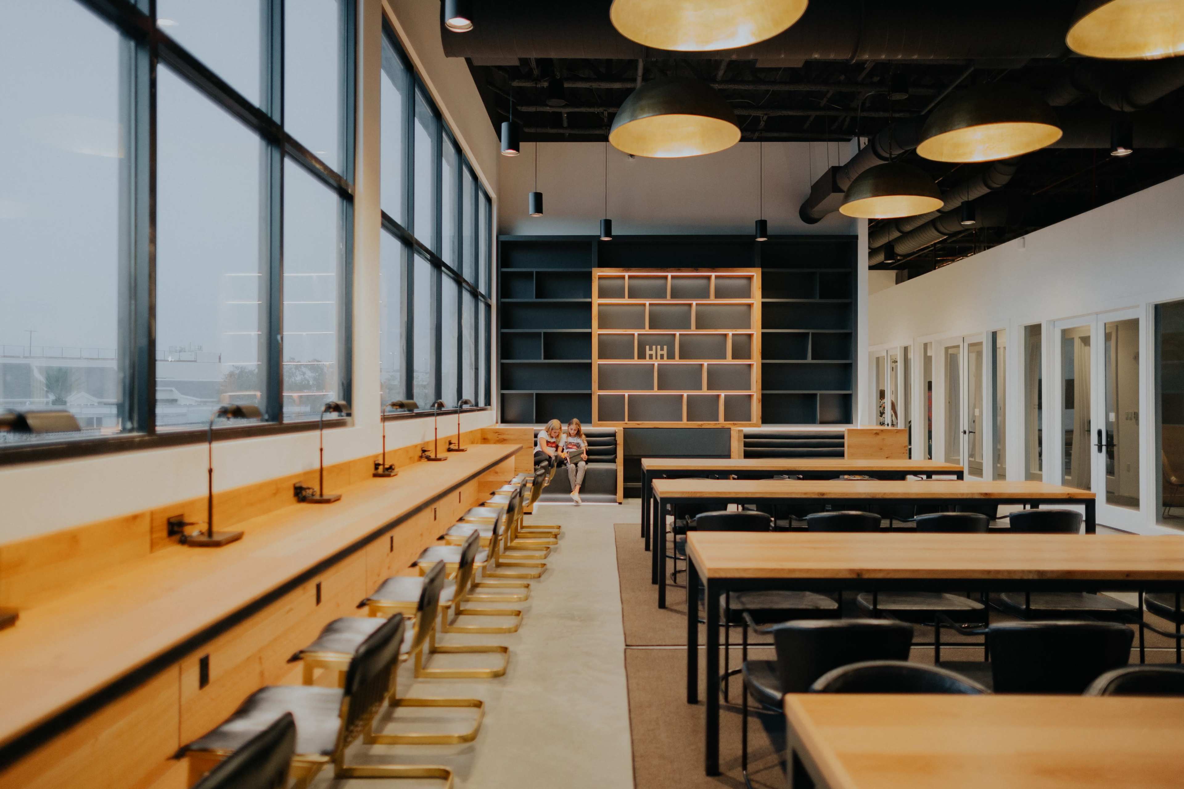 A modern, spacious office interior features long wooden tables, black chairs, large windows, and a bookshelf against the back wall.