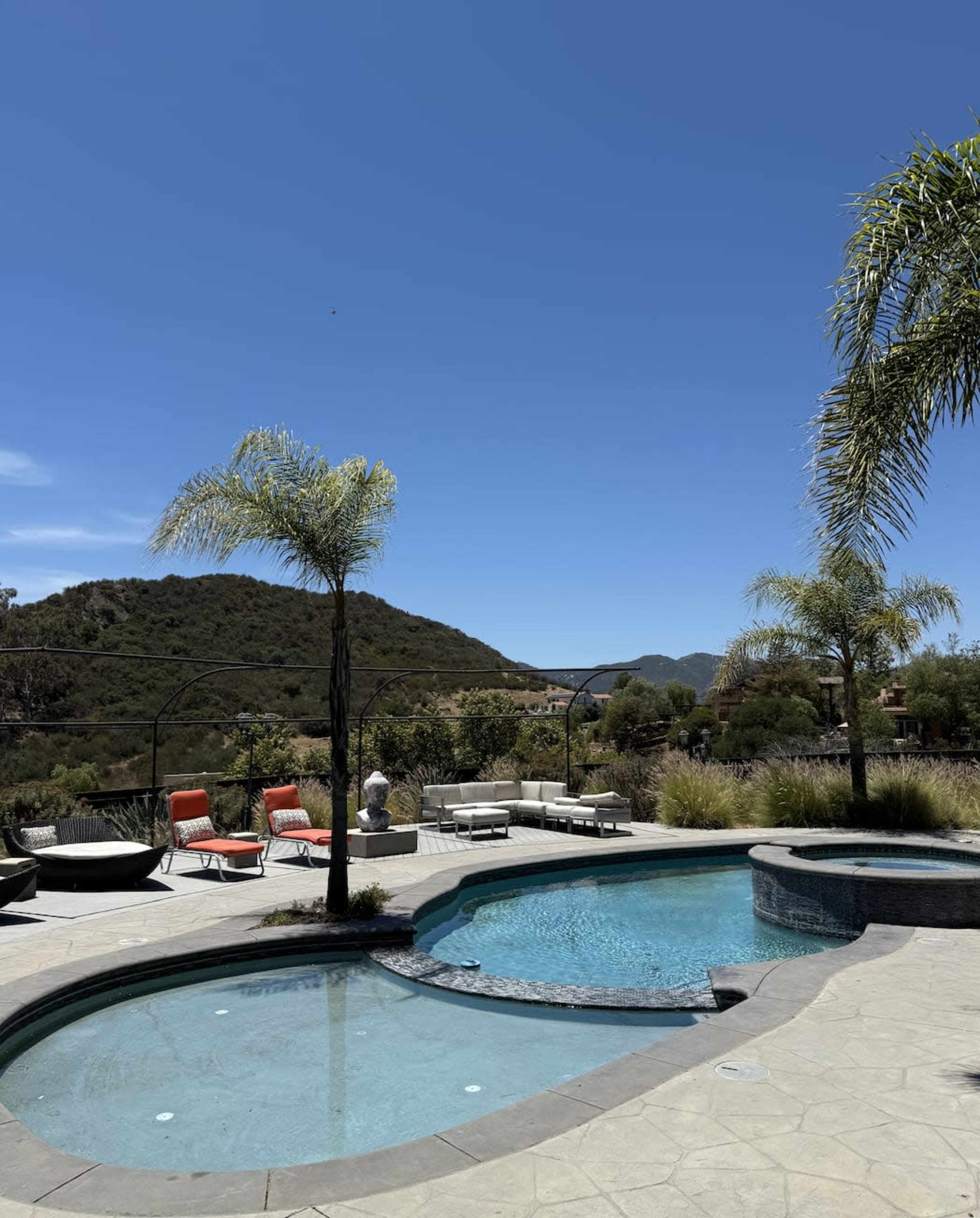 The image depicts a backyard with a swimming pool surrounded by lounge chairs and palm trees, set against a backdrop of hills under a clear blue sky.