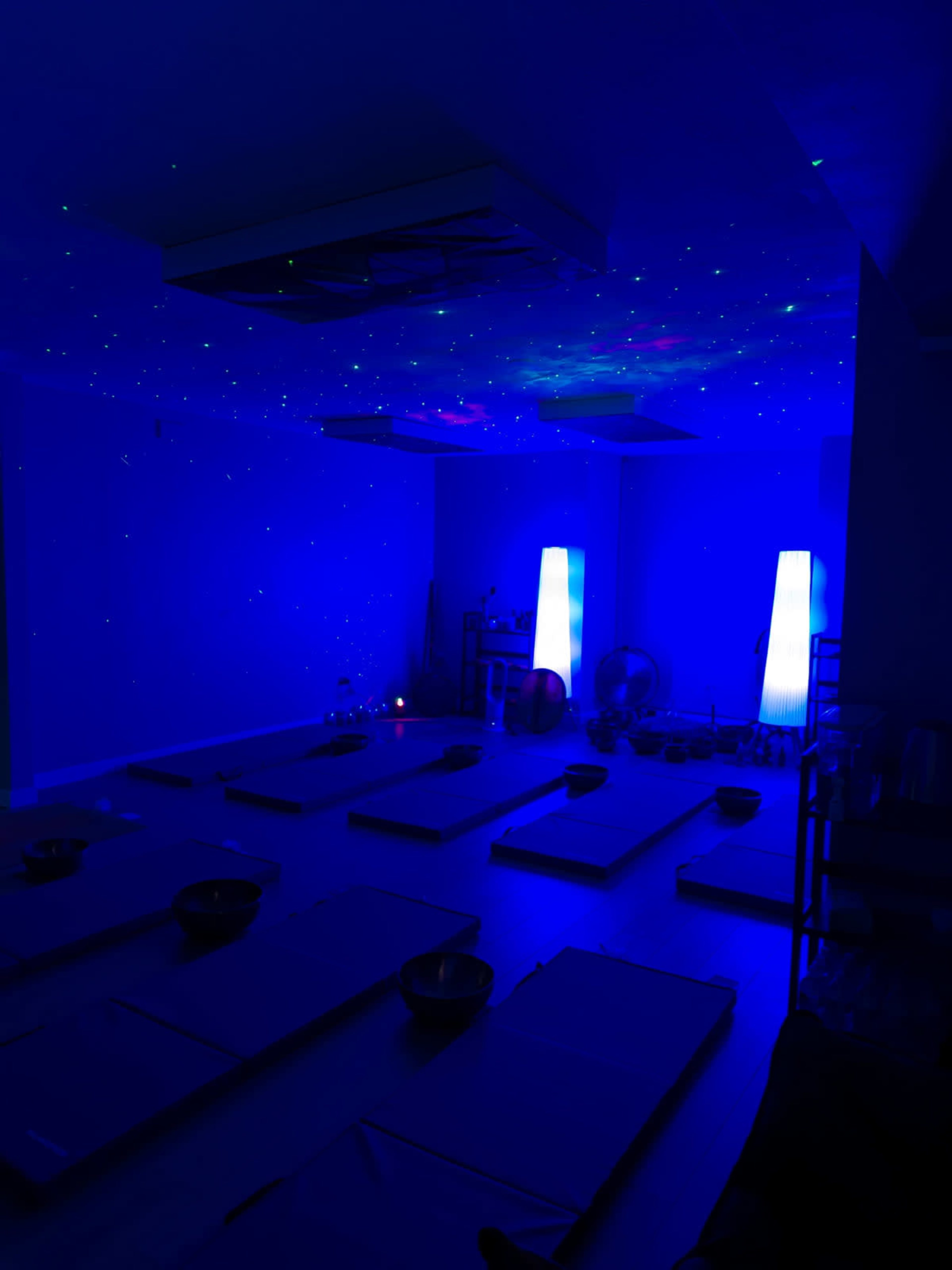 The image shows a dimly lit meditation room with blue lighting, featuring several yoga mats arranged on the floor and two illuminated column lights.