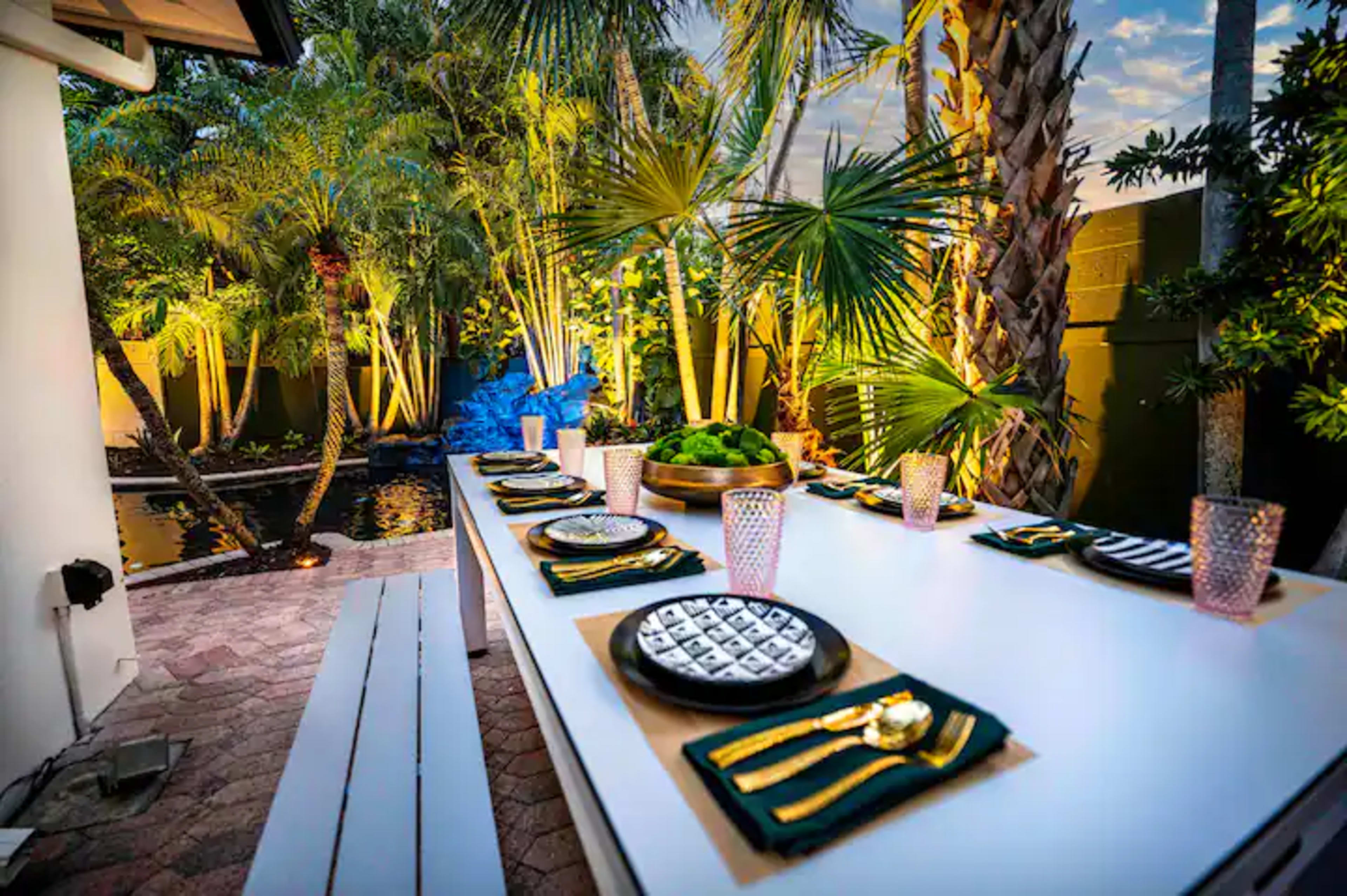 An outdoor dining table set with plates, glasses, and silverware, surrounded by lush greenery and illuminated by soft lighting.