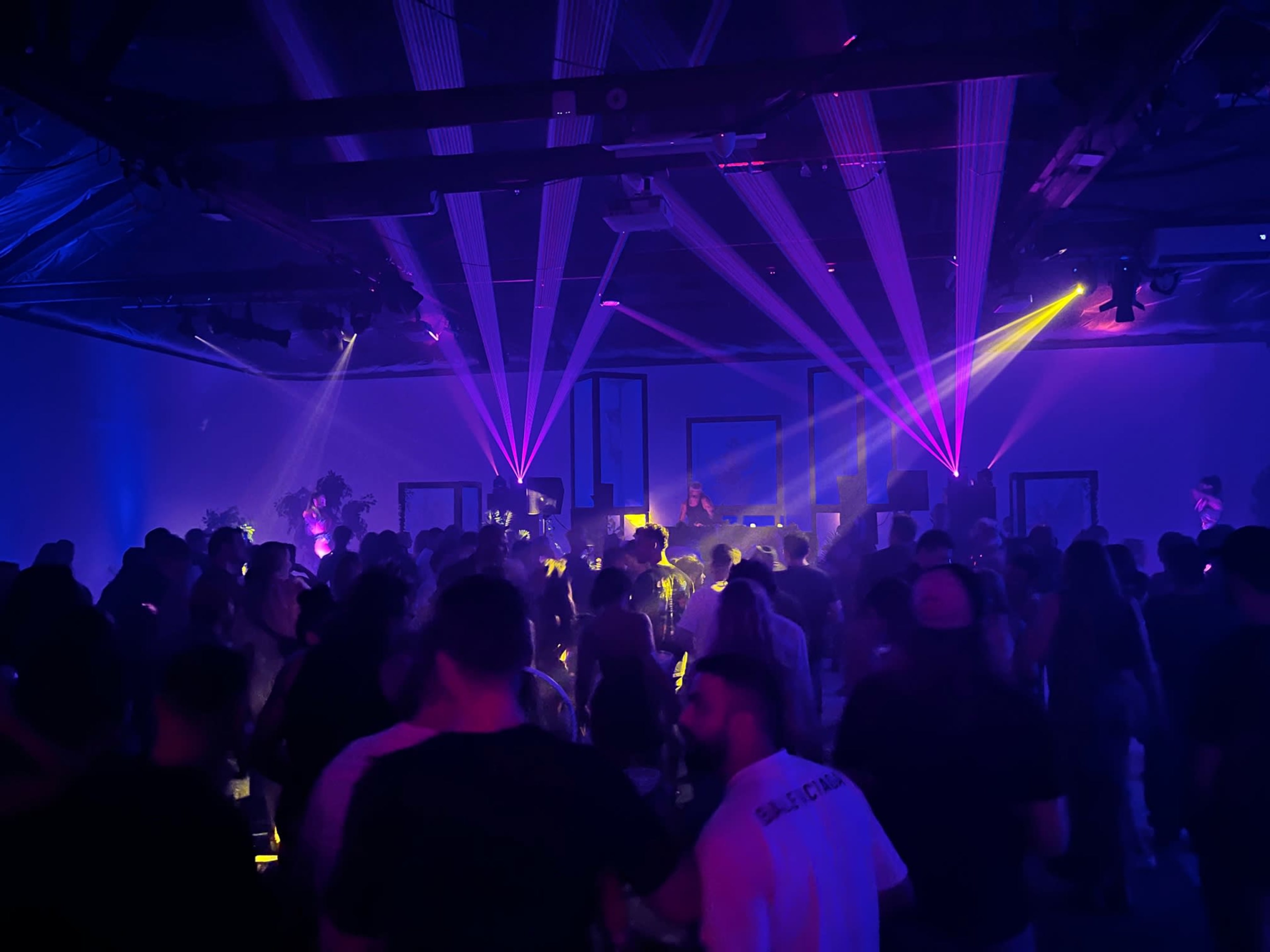 A crowded indoor venue features people dancing under colorful laser lights.